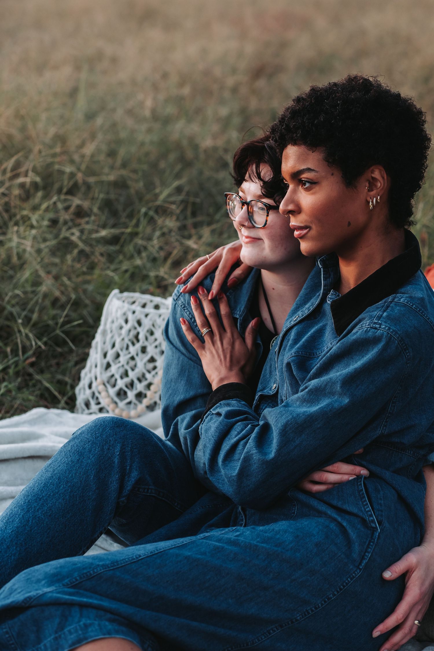 Two women in denim sit together in a field, one embracing the other from behind, sharing a tender moment.