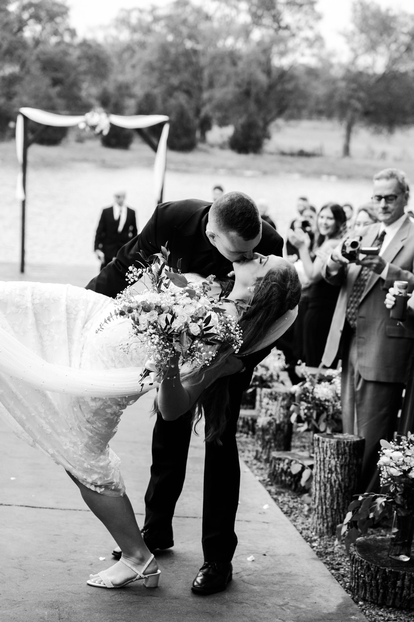 Black and white photo of groom dipping bride for a kiss at outdoor wedding ceremony with guests watching.