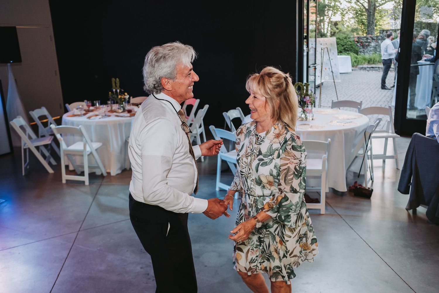 A couple dances together during a reception in an elegant event space with white chairs and natural lighting.