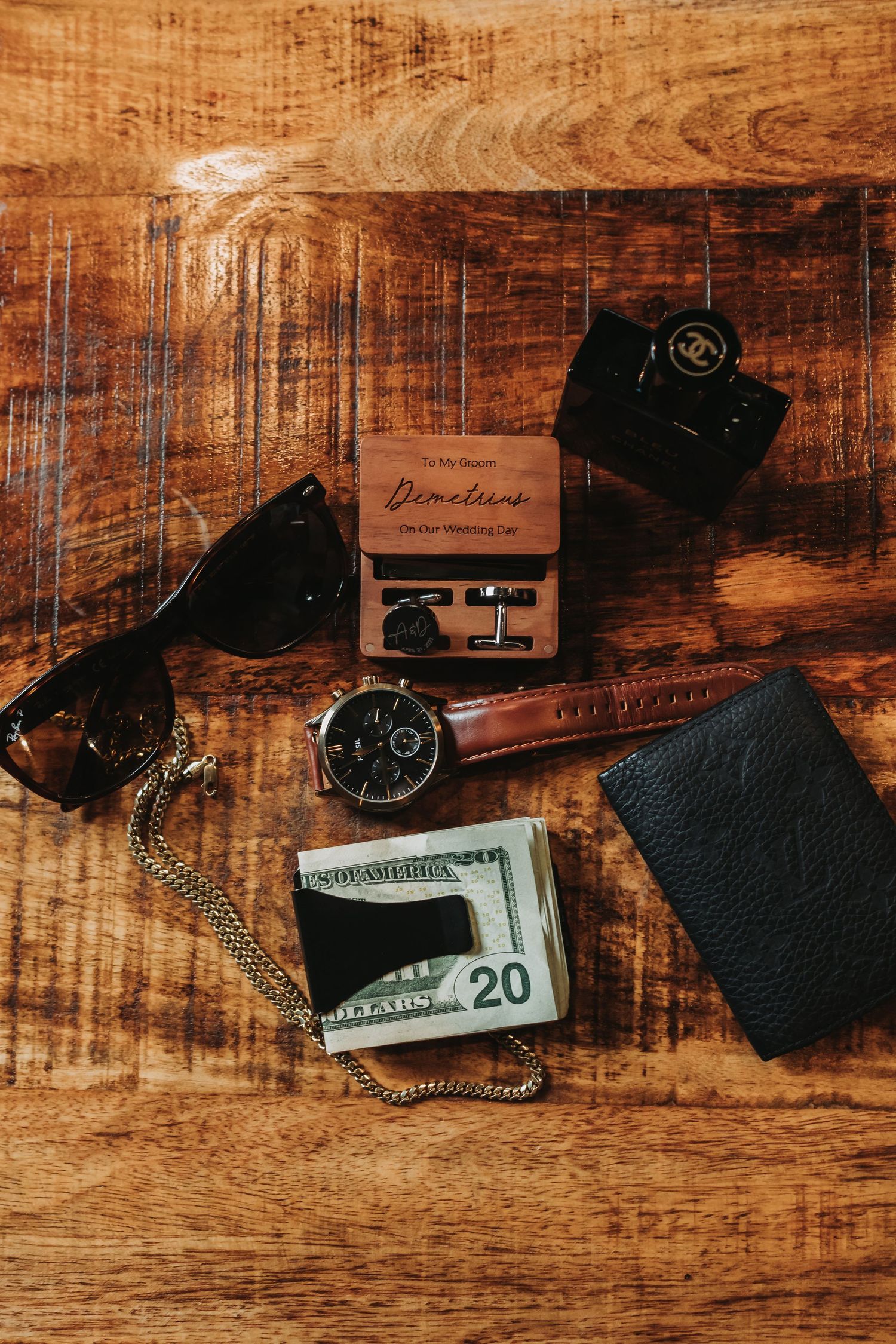 Everyday men's accessories including sunglasses, wallet, watch, money clip and lighter arranged on rustic wooden surface.