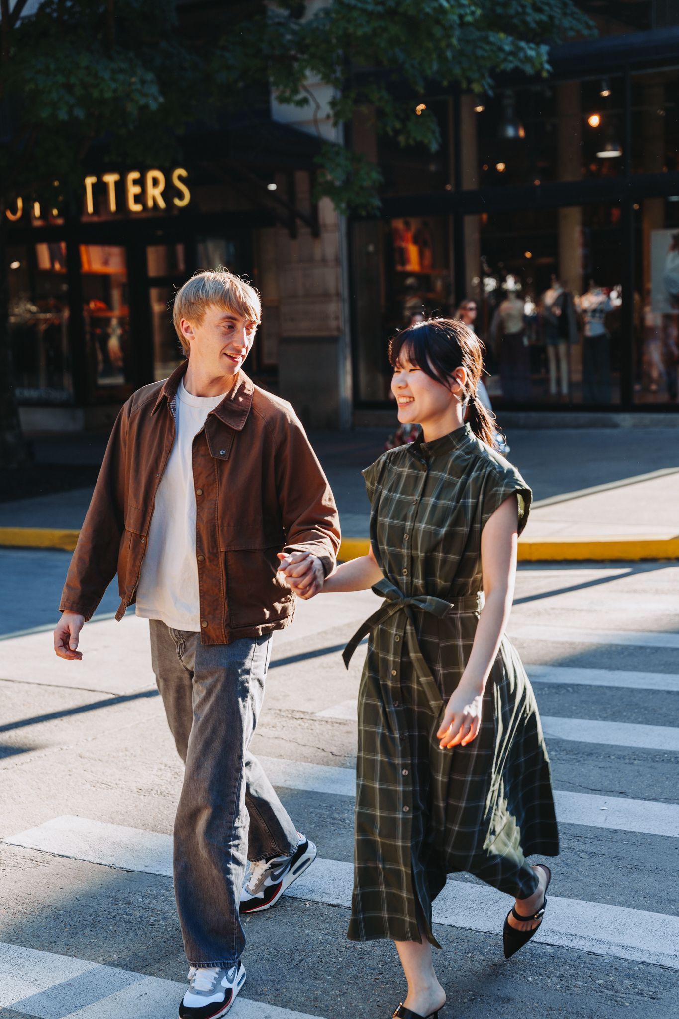 Young couple holding hands crossing a city street, man in brown jacket and woman in green plaid dress, smiling.