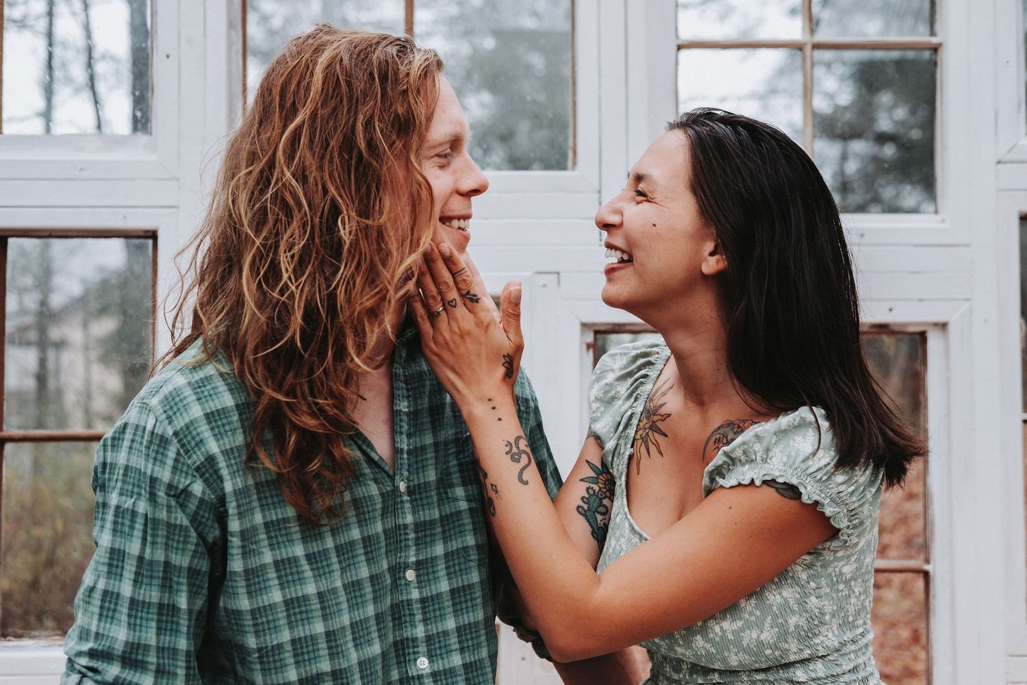 A smiling couple shares a tender moment, woman with tattoos touching mans face near white window.