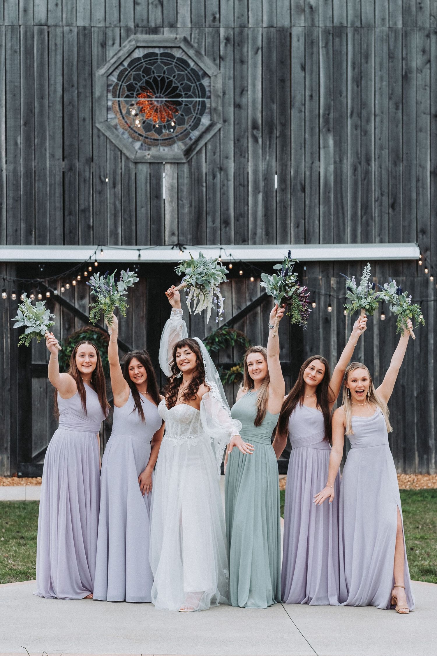 A group of bridesmaids in lavender and sage dresses celebrating with bouquets in front of a rustic barn wall.