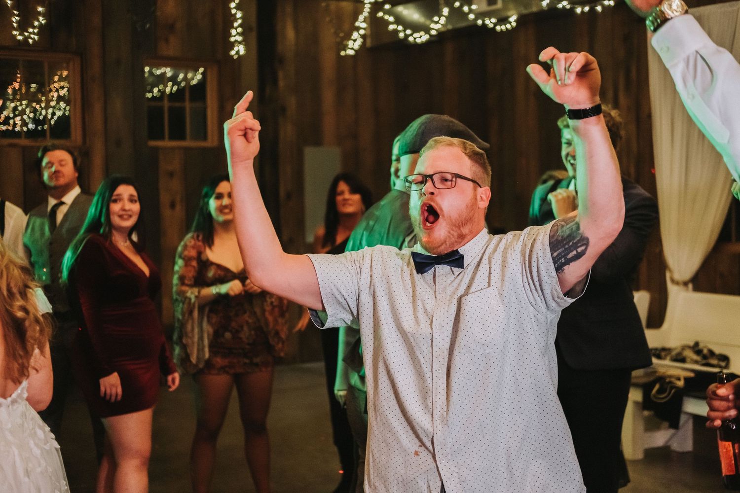 An enthusiastic wedding guest dancing with raised arms in casual attire at a reception with twinkle lights behind them.