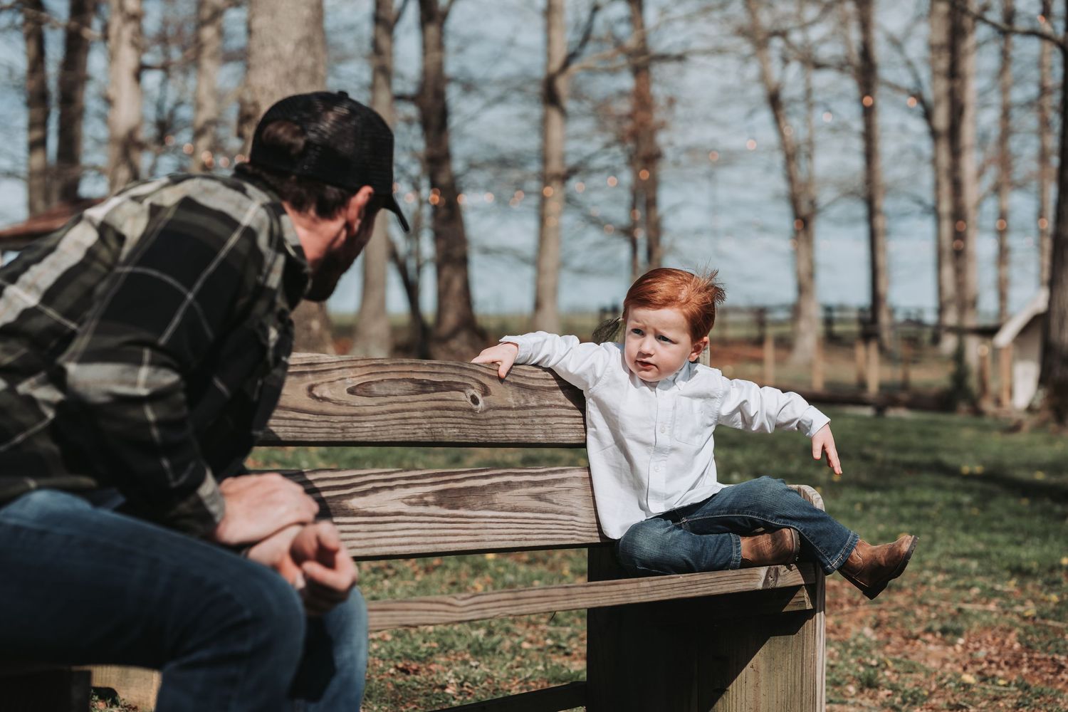A child balances playfully on a wooden fence while engaging with someone in a plaid shirt on an autumn day.