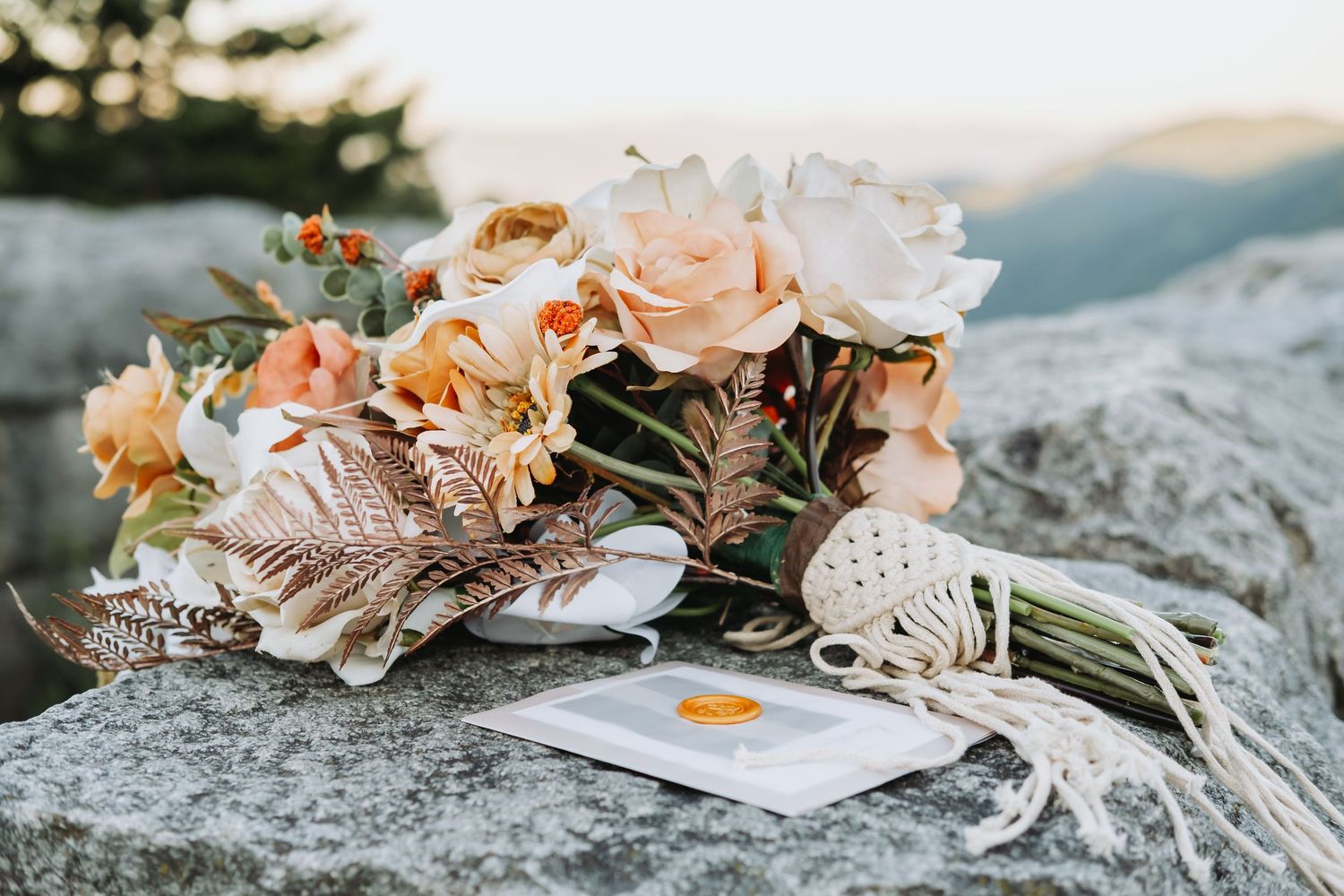 A bouquet of peach roses and dried flowers laid on a rock with a small note card at sunset in the mountains.