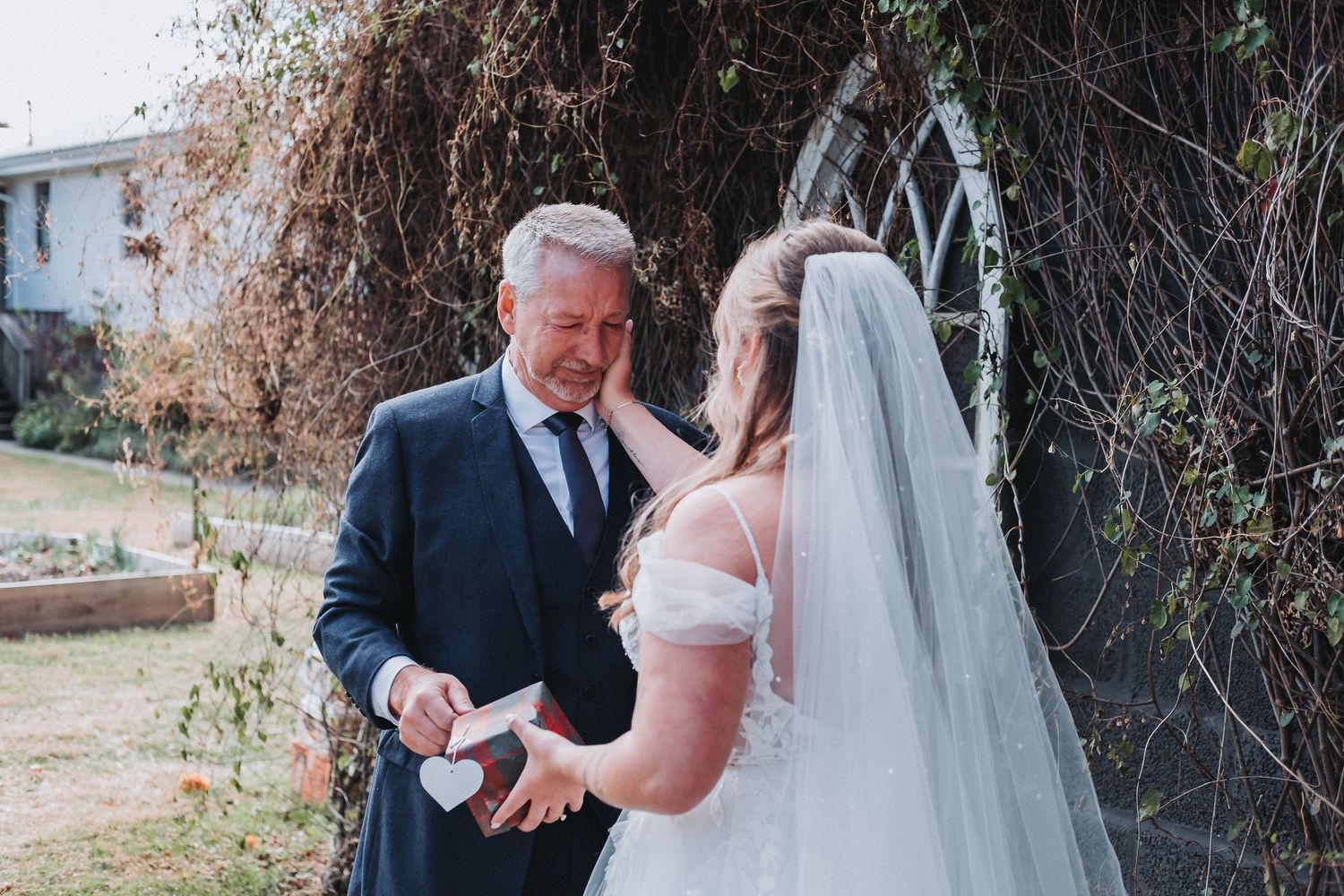 A bride in a white dress and long veil shares an emotional outdoor moment next to a decorative garden arch.