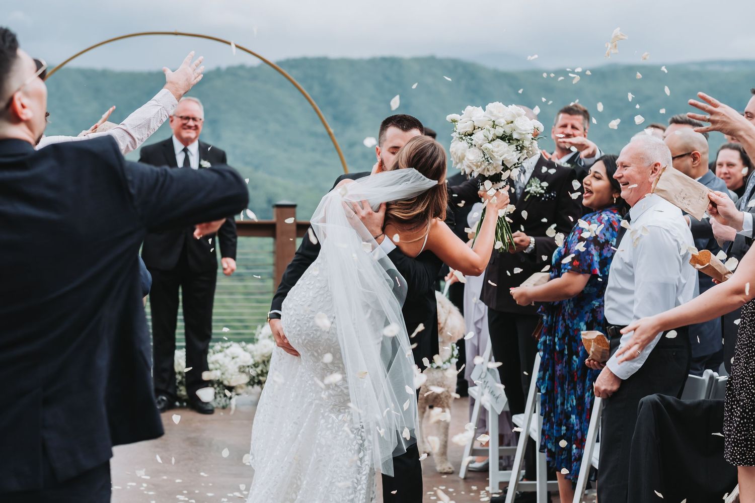 Wedding party celebrates under floral arch at lakeside ceremony with guests throwing confetti and flower petals.