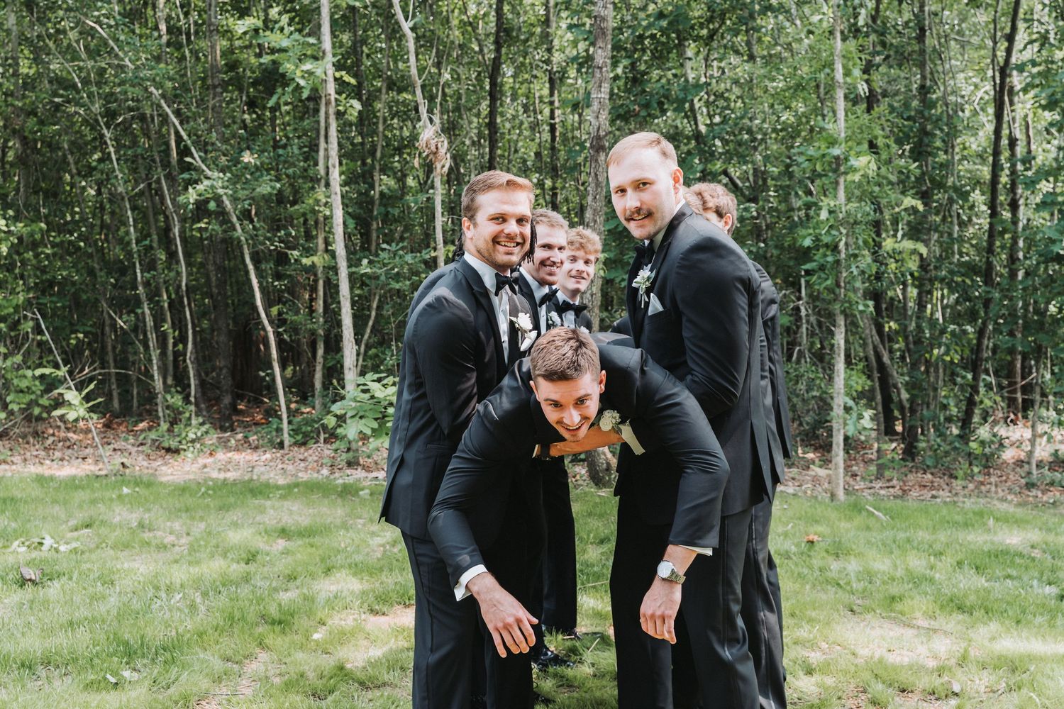 Two men in black suits stand together outdoors in a wooded area sharing an emotional moment.