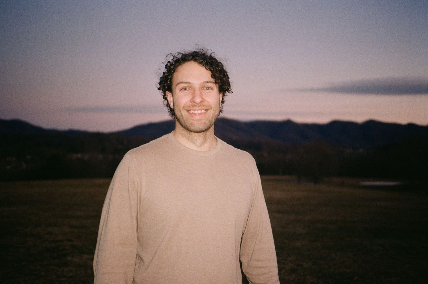 Smiling young man with curly hair in beige sweater standing outdoors at dusk with mountain backdrop.
