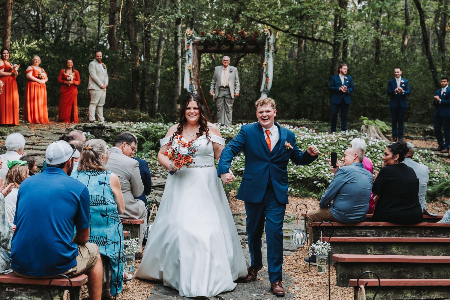 Guests seated on wooden benches during an outdoor garden wedding ceremony in a wooded setting at dusk.