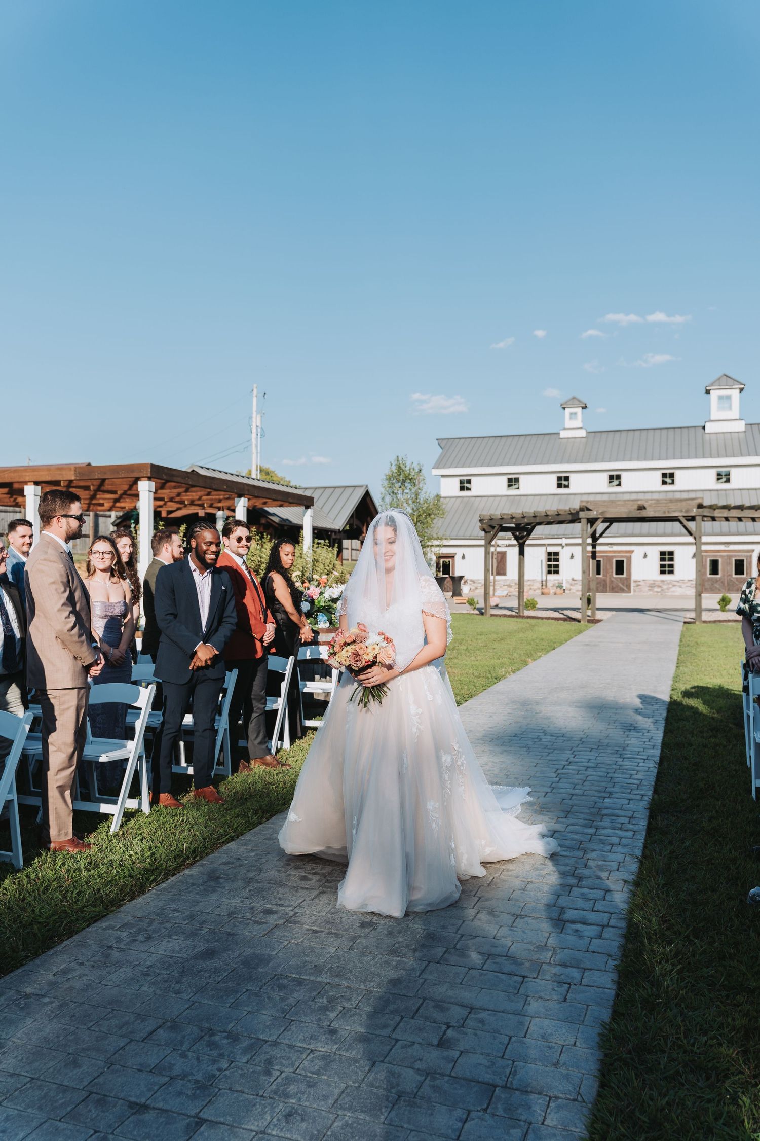 Wedding party gathers outdoors on a stone pathway with white buildings and blue sky in the background.
