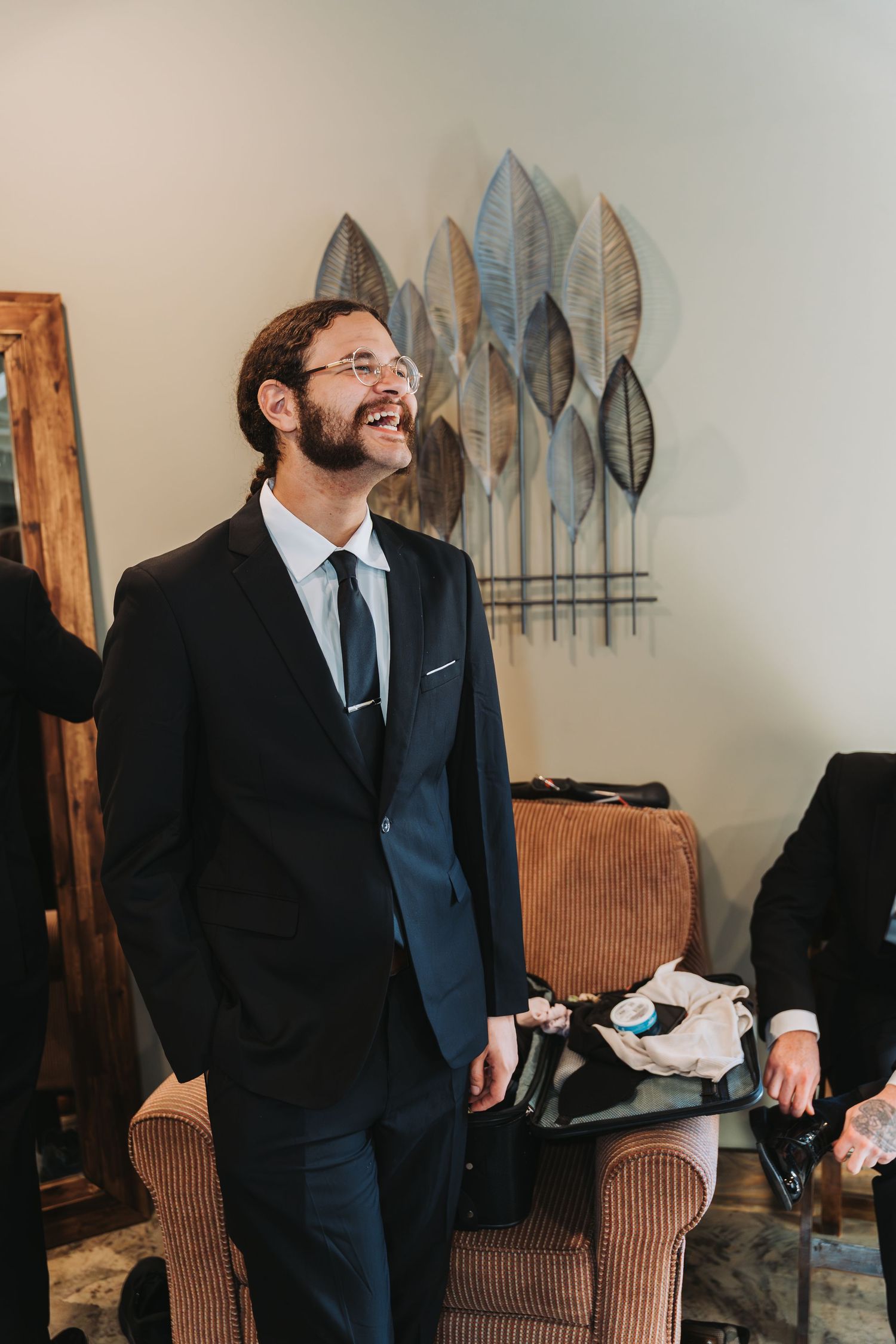 A person in a dark suit laughs heartily while sitting on a brown armchair in a room with decorative wall art.
