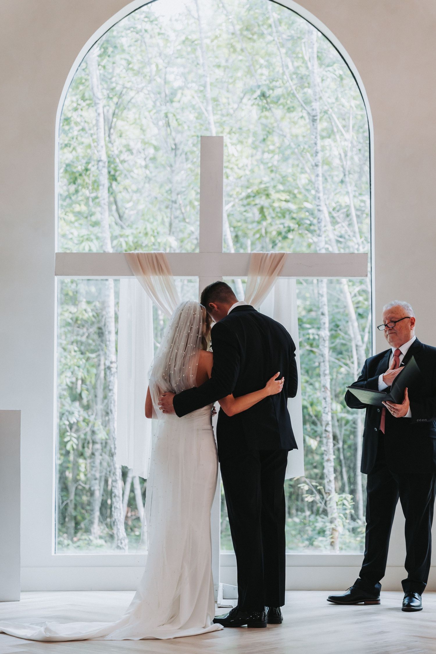 A couple shares their first kiss during a wedding ceremony in front of a large arched window with natural lighting.