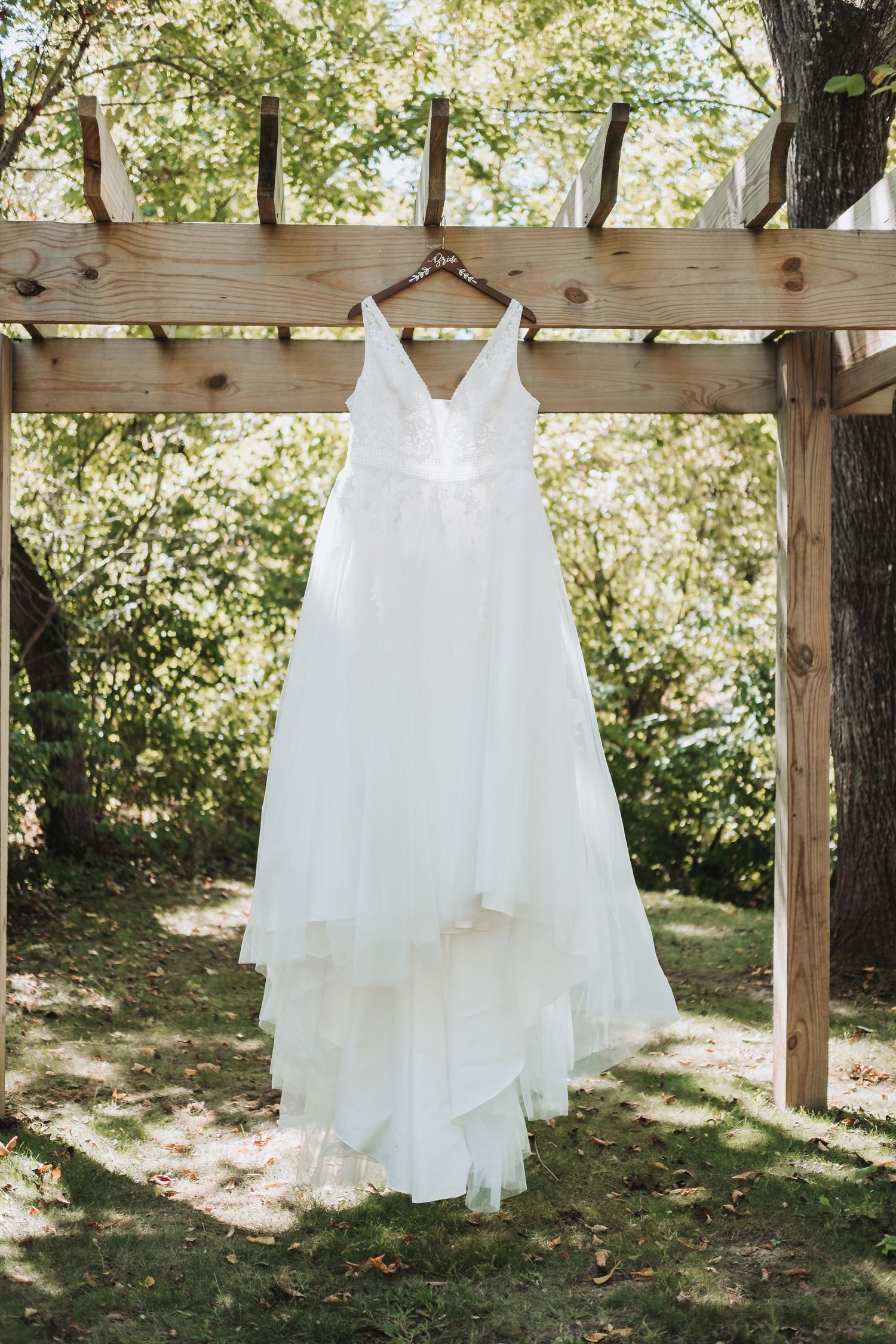 A white wedding dress hangs elegantly from a wooden pergola in a sunlit garden setting.