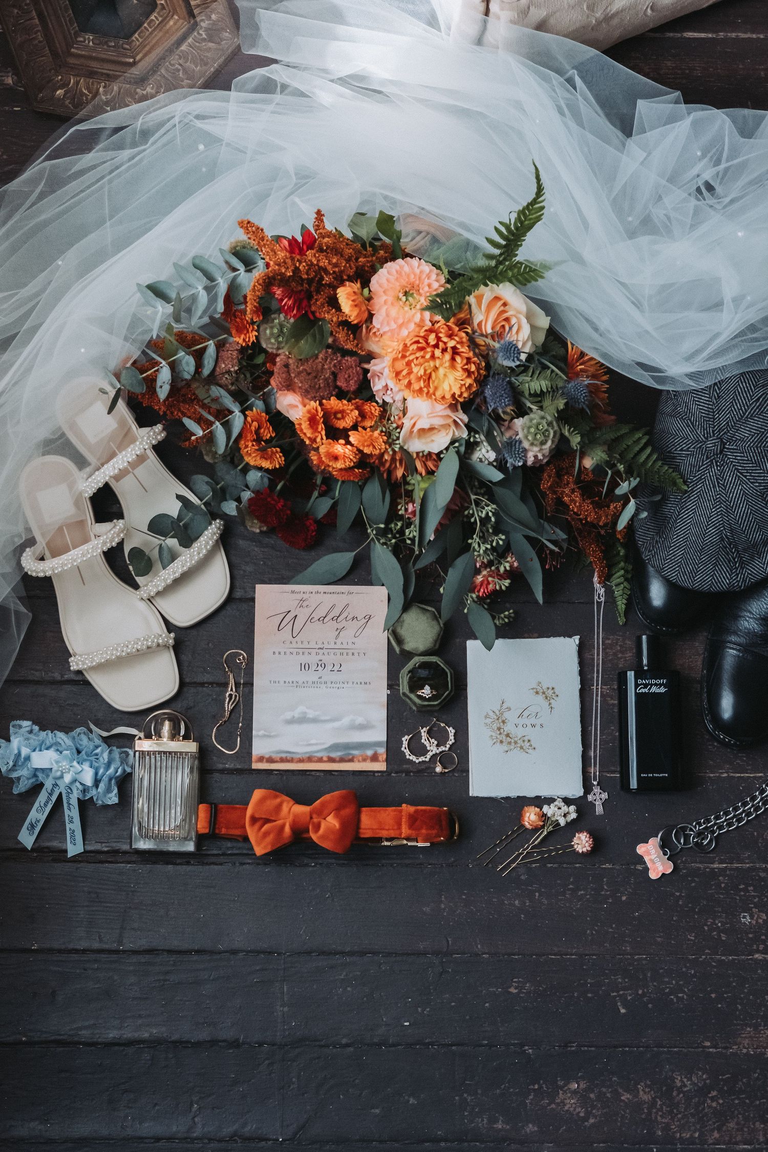 Wedding details flatlay with peach and coral flowers, white tulle veil, rings, stationery and orange bow tie on dark wood surface.