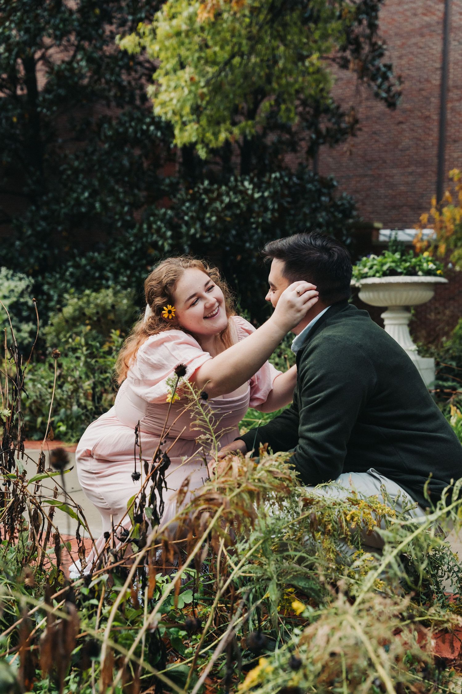 A couple shares a playful moment together in a lush garden setting with brick buildings in the background.