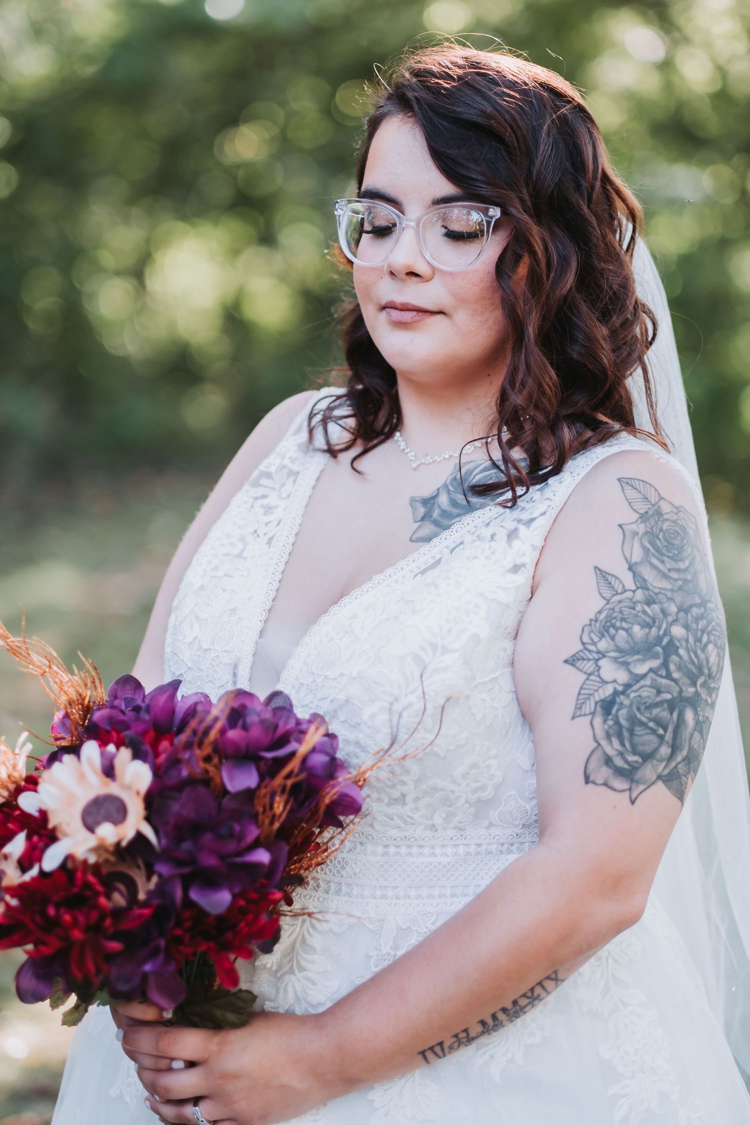 Beautiful bride in lace wedding dress holding vibrant purple and burgundy bouquet, with floral tattoos and glasses in sunlit setting.