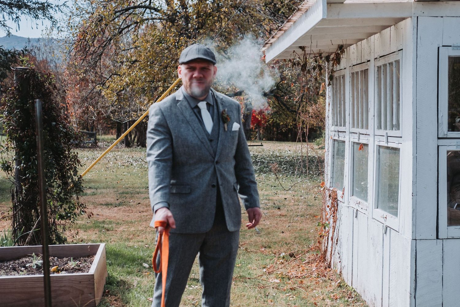 Well-dressed person in a gray suit jacket standing outdoors next to a white building with visible smoke in autumn setting.