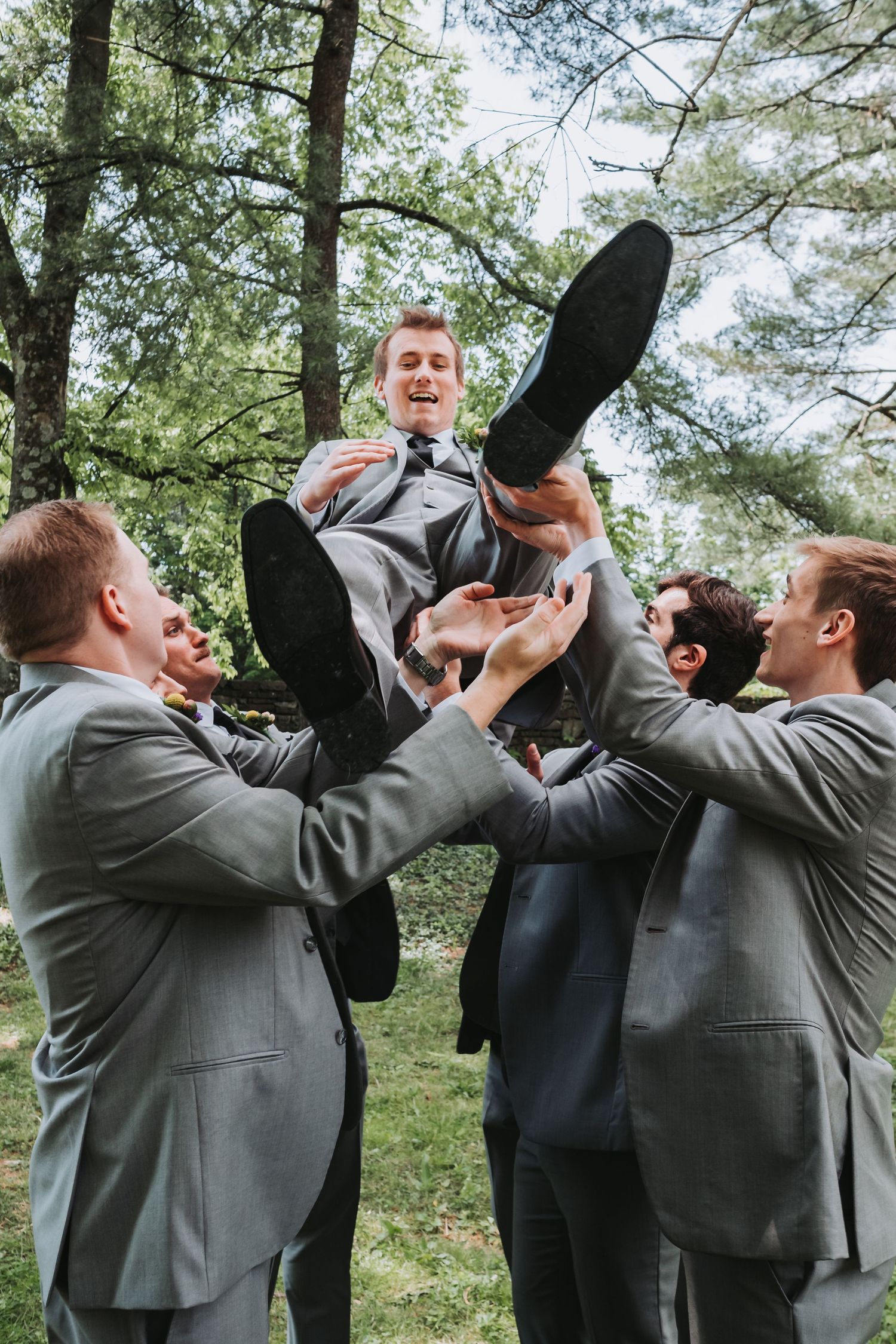 Four groomsmen in gray suits perform a celebratory champagne toast together outdoors beneath tall pine trees.