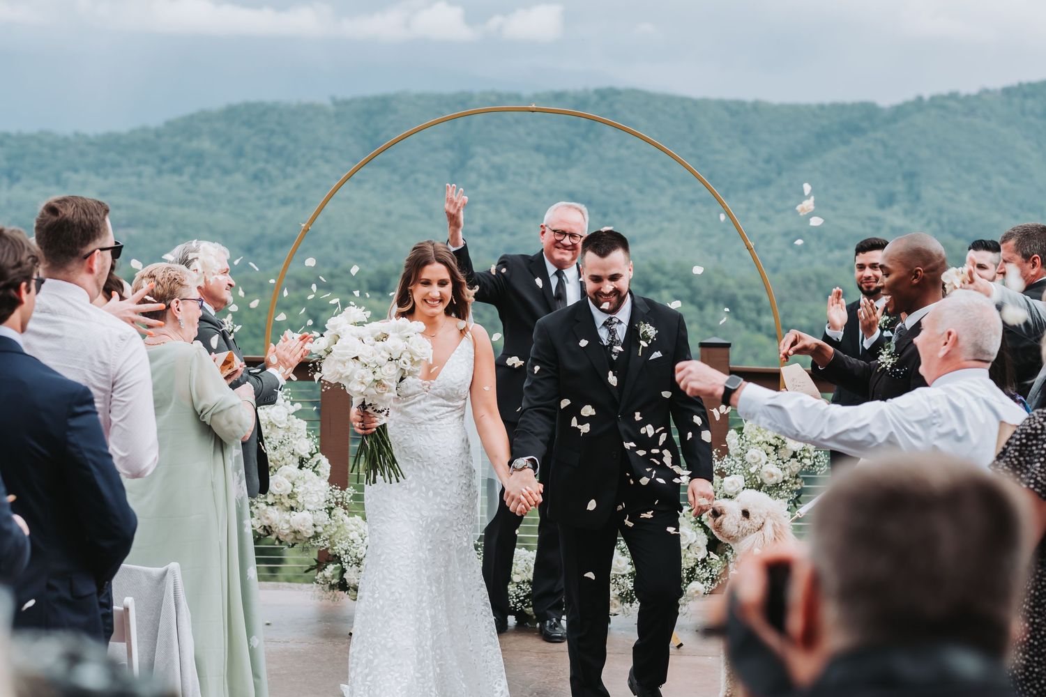 Newlyweds celebrate under a golden arch while guests shower them with confetti in a mountain setting.