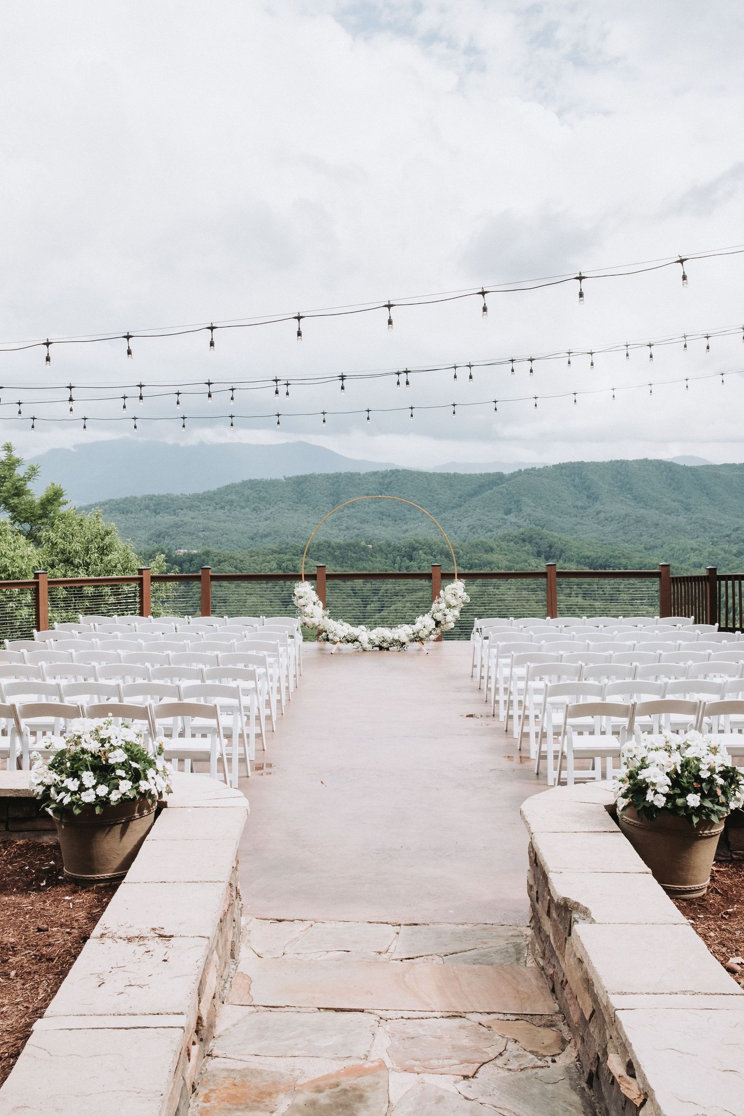 Mountain wedding ceremony setup with white chairs, string lights and floral decorations on an outdoor stone terrace.