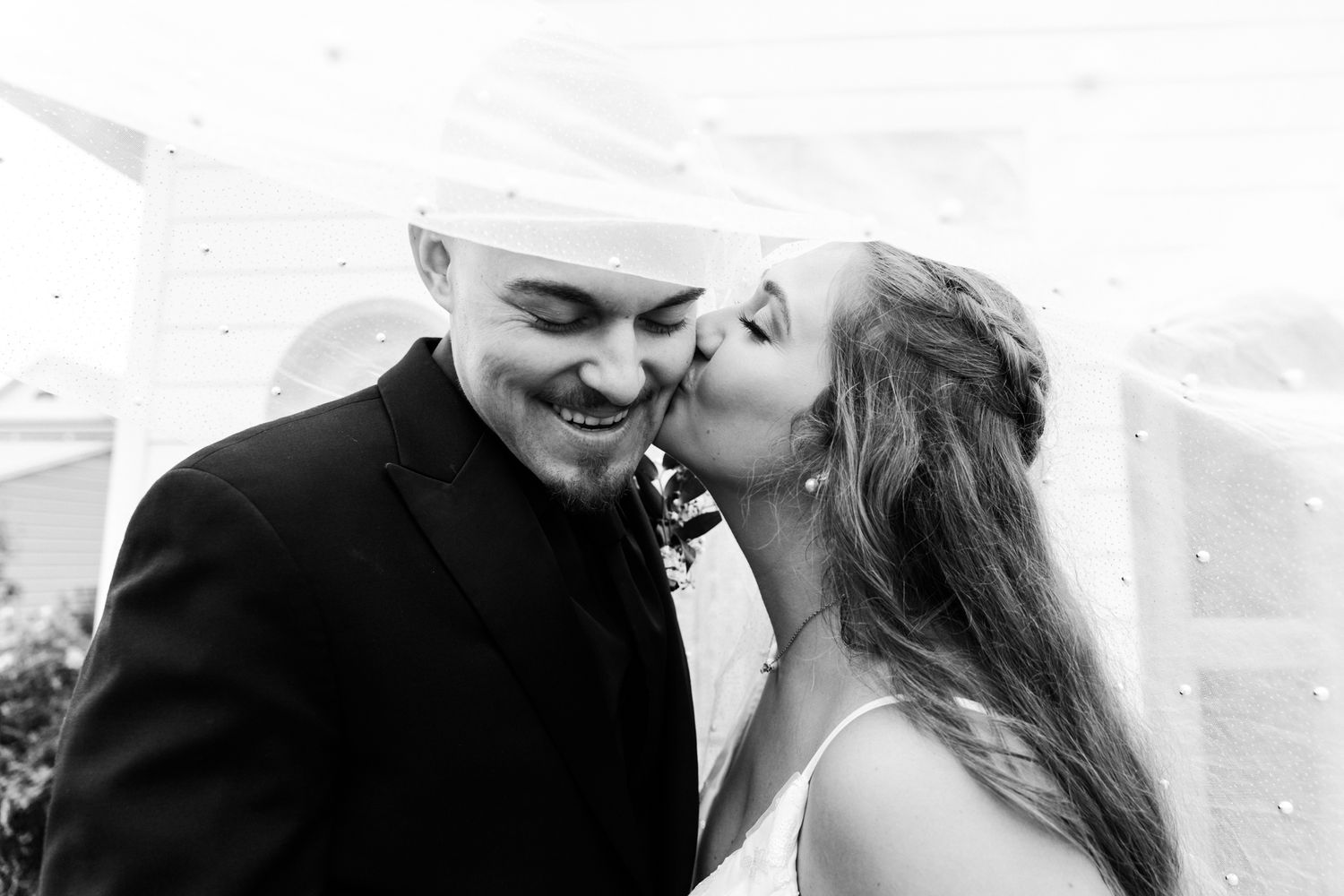 Black and white photo of a bride kissing groom on cheek, both smiling, veil flowing, wedding day romance.
