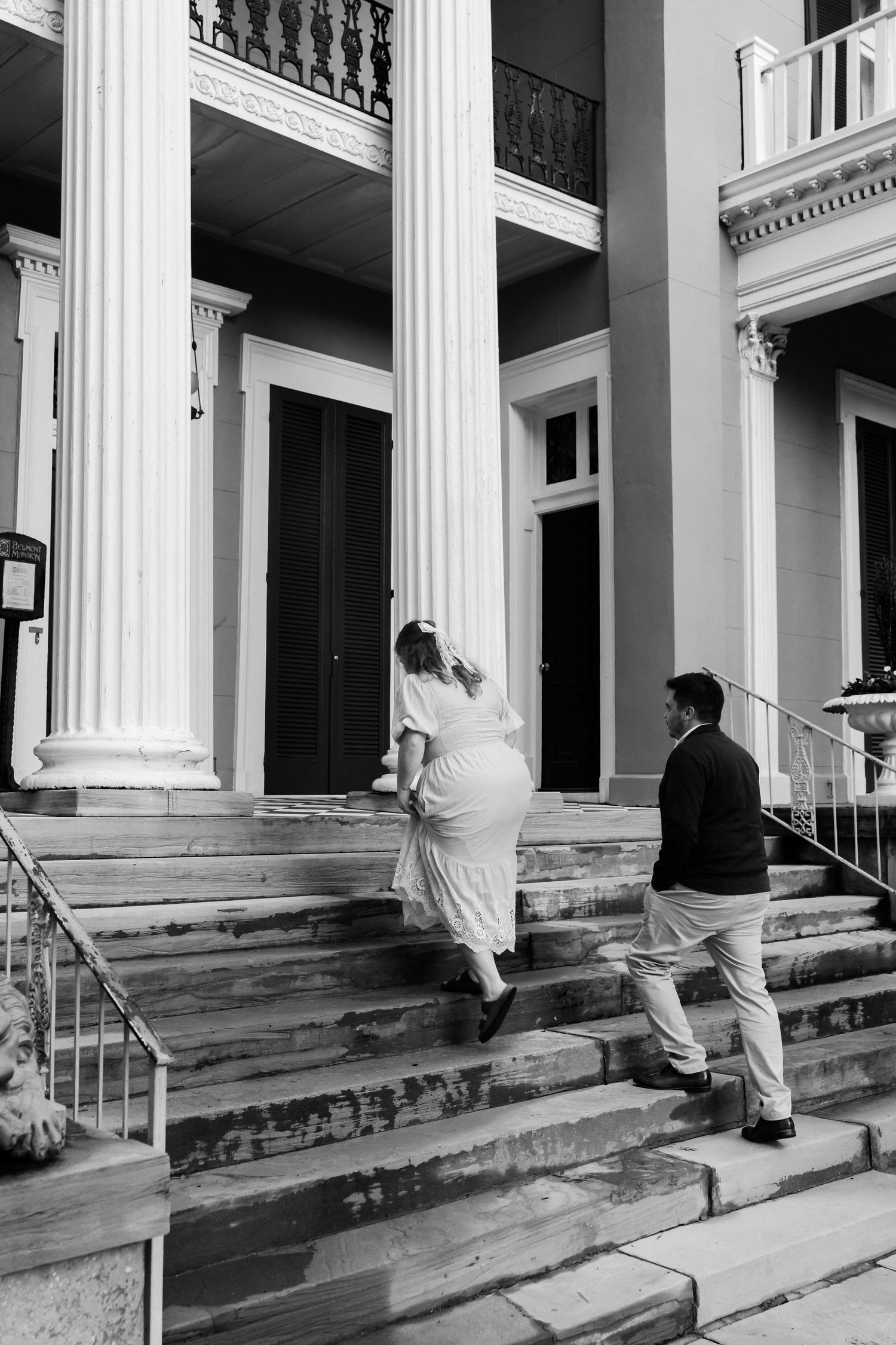 A couple ascends the grand steps of a historic building with white columns and ornate balconies in black and white.