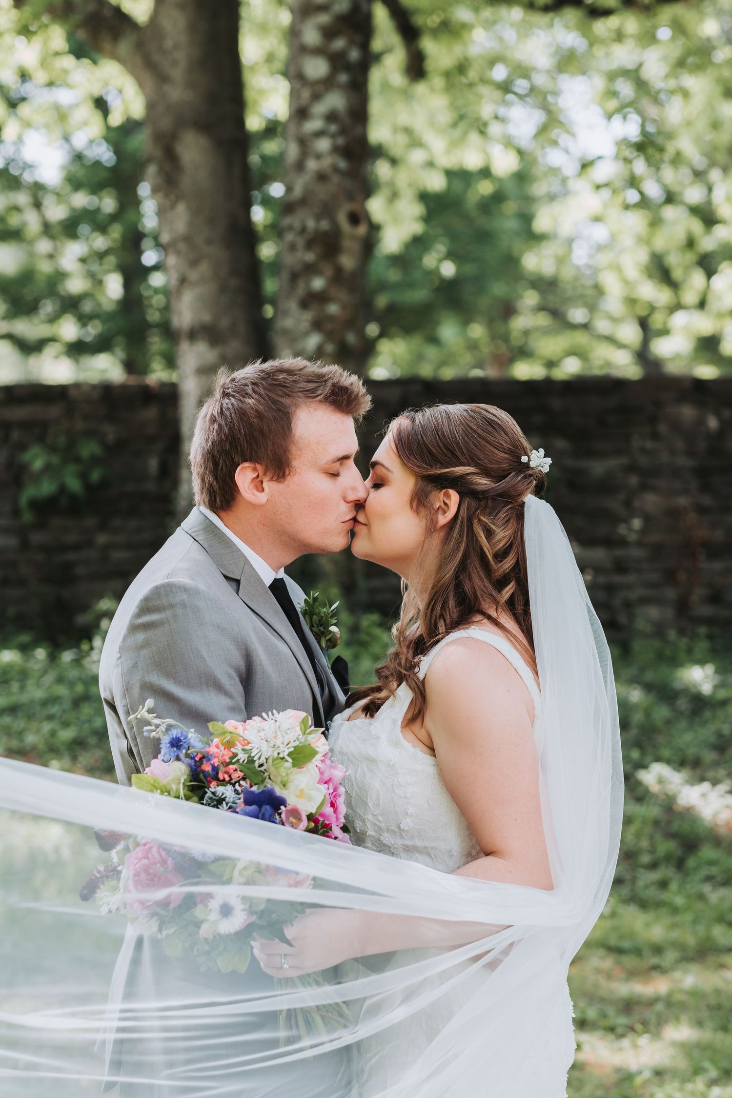 Newlyweds share a romantic kiss at their outdoor wedding with blooming wildflowers and a flowing veil in sunlit garden.