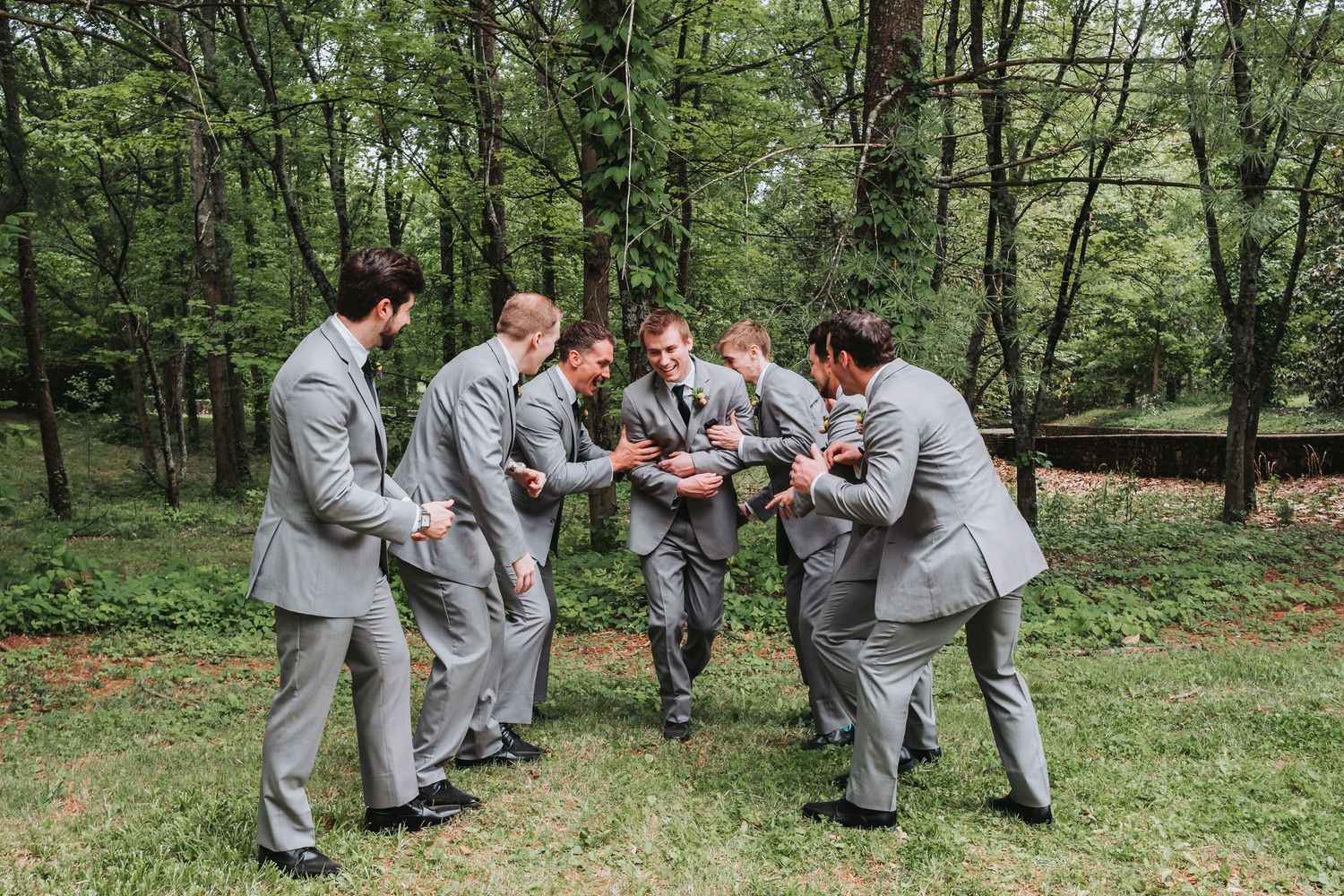 A group in matching gray suits gather in a playful formation outdoors surrounded by trees in a wooded area.