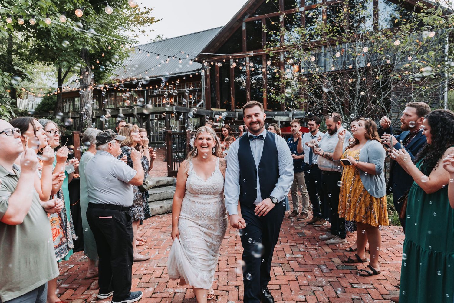 Wedding guests dancing outdoors on brick patio during sunset reception at rustic venue with string lights overhead.