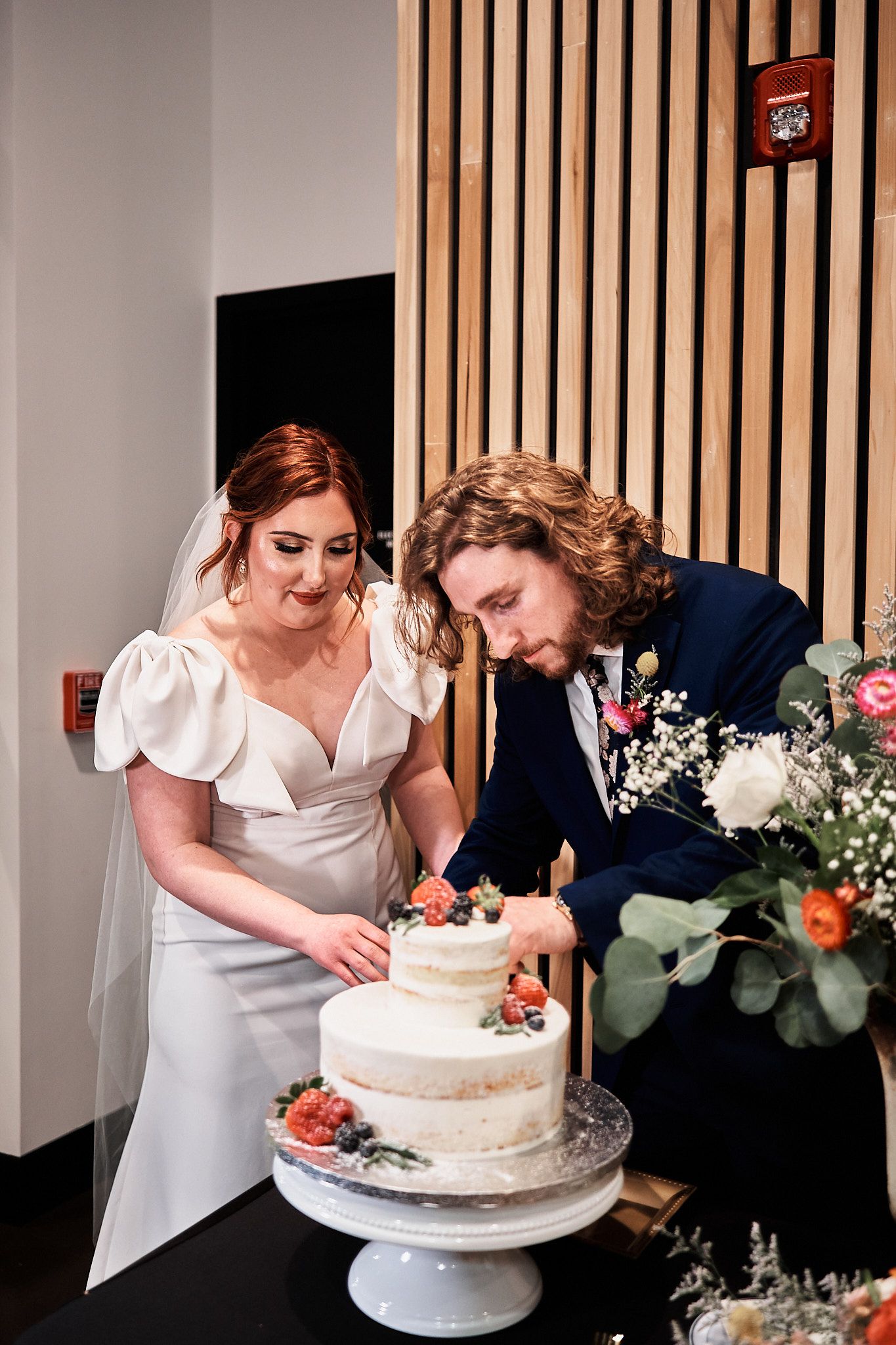 Newlyweds in white dress and navy suit cut their rustic wedding cake decorated with fresh flowers and berries.
