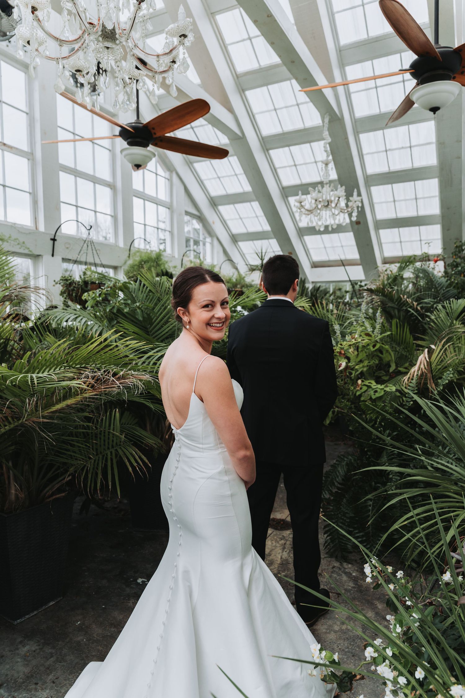 Elegant bride in white mermaid gown glances back while standing in a greenhouse conservatory with tall windows and chandeliers.
