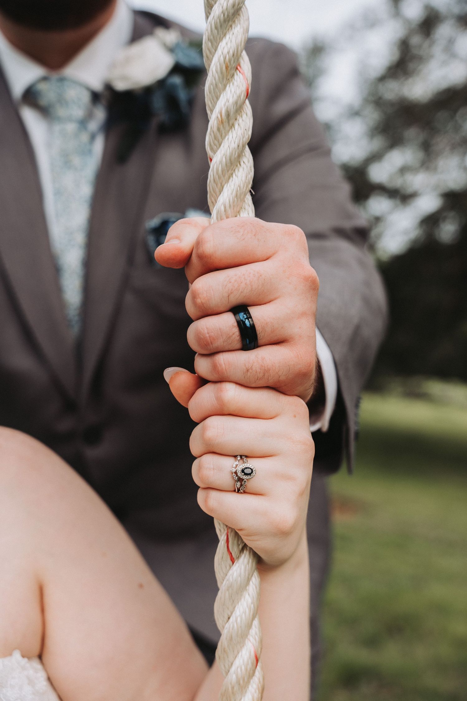 Close-up shot of hands with wedding rings holding a thick white rope outdoors against a natural background.