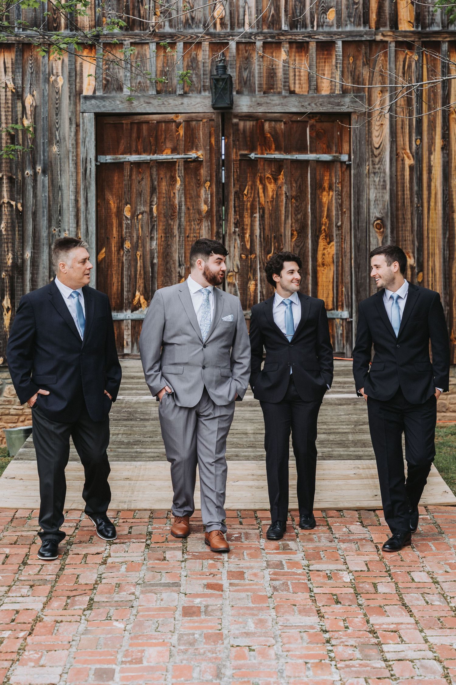 Groomsmen in formal suits pose casually against a rustic wooden wall on brick pavement in a series of candid shots.