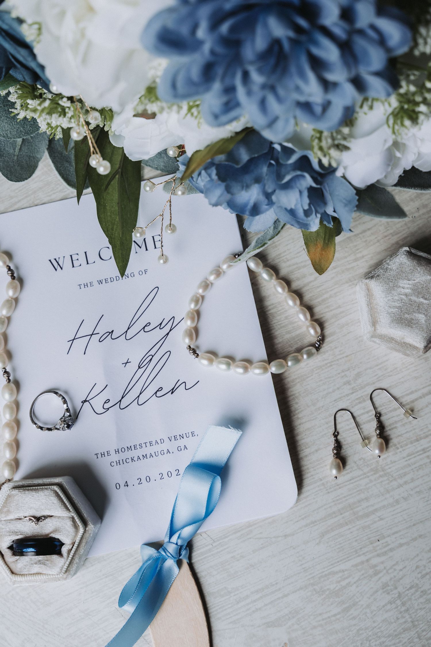 Elegant wedding invitation with blue flowers and delicate pearl accessories on a light wooden surface.
