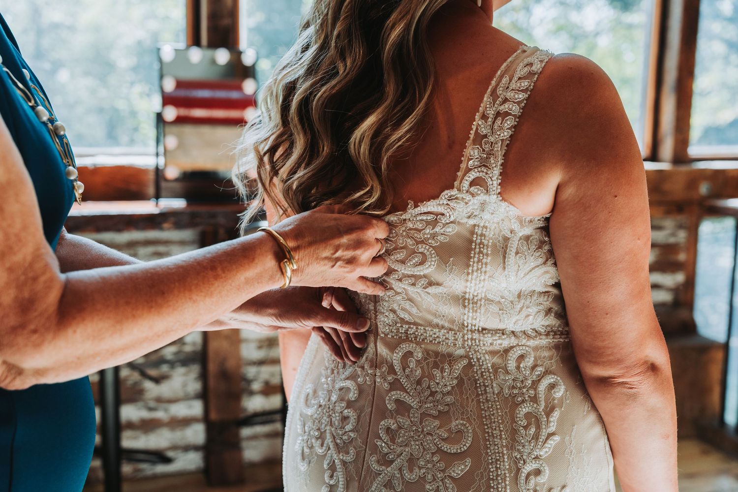 A bride in a lace wedding dress gets help fastening her gown from a helper in a blue dress indoors.