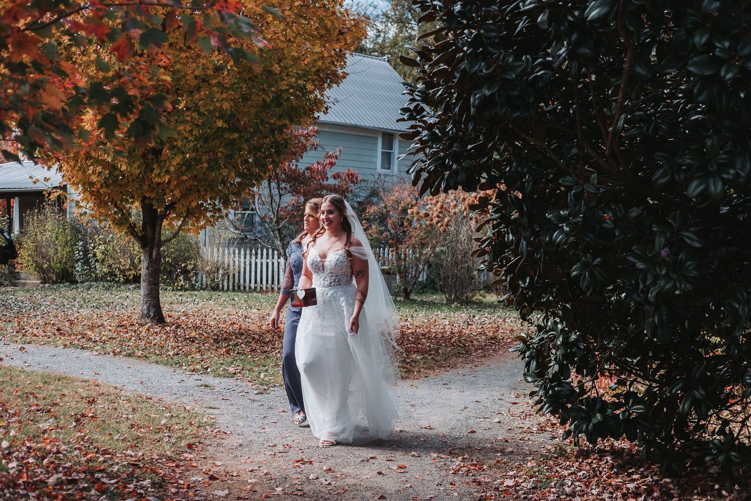 A couple in autumn attire walks along a tree-lined path with orange and red fallen leaves on the ground.