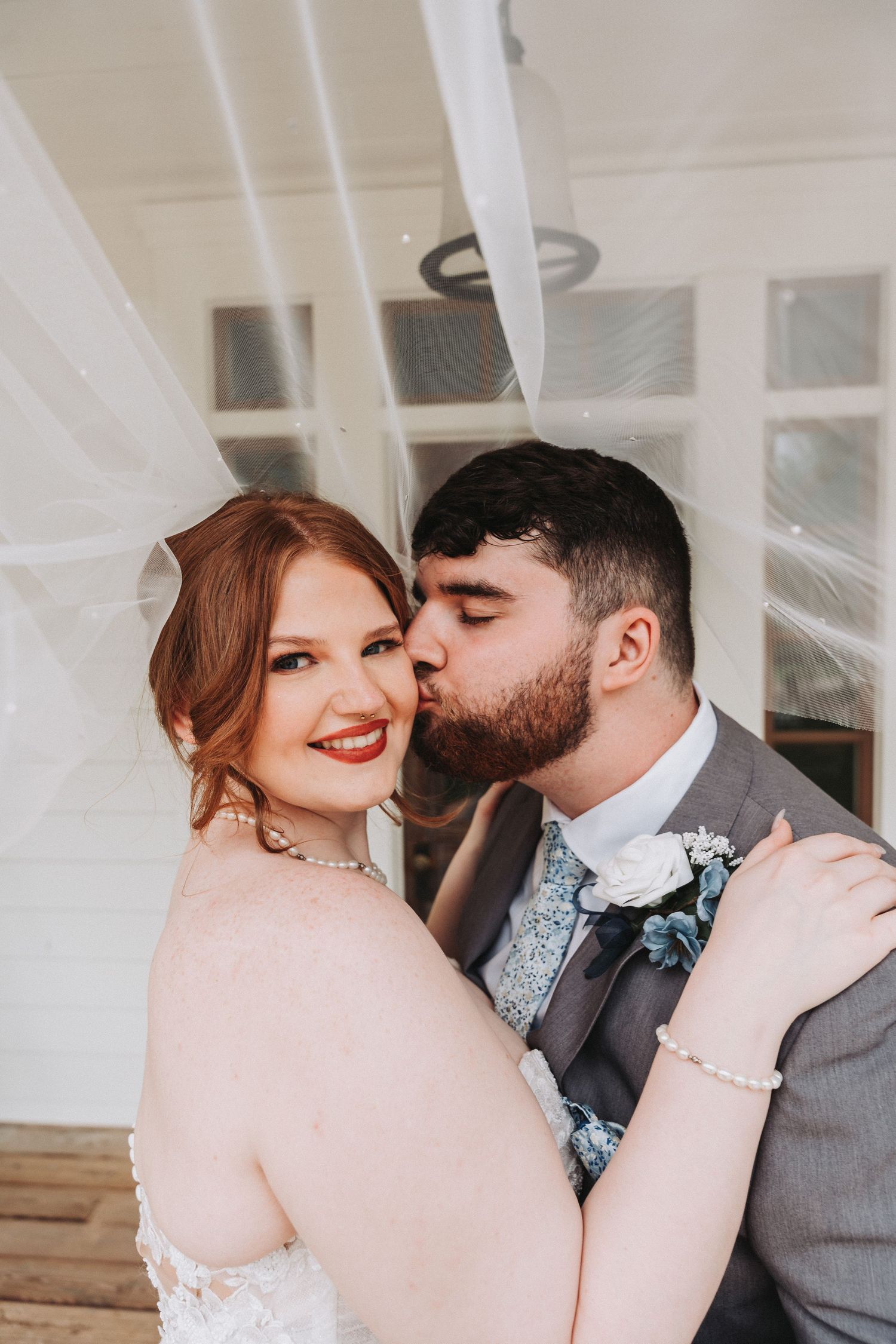 A romantic wedding portrait with a couple embracing under sheer white veils while wearing blush pink and gray attire.