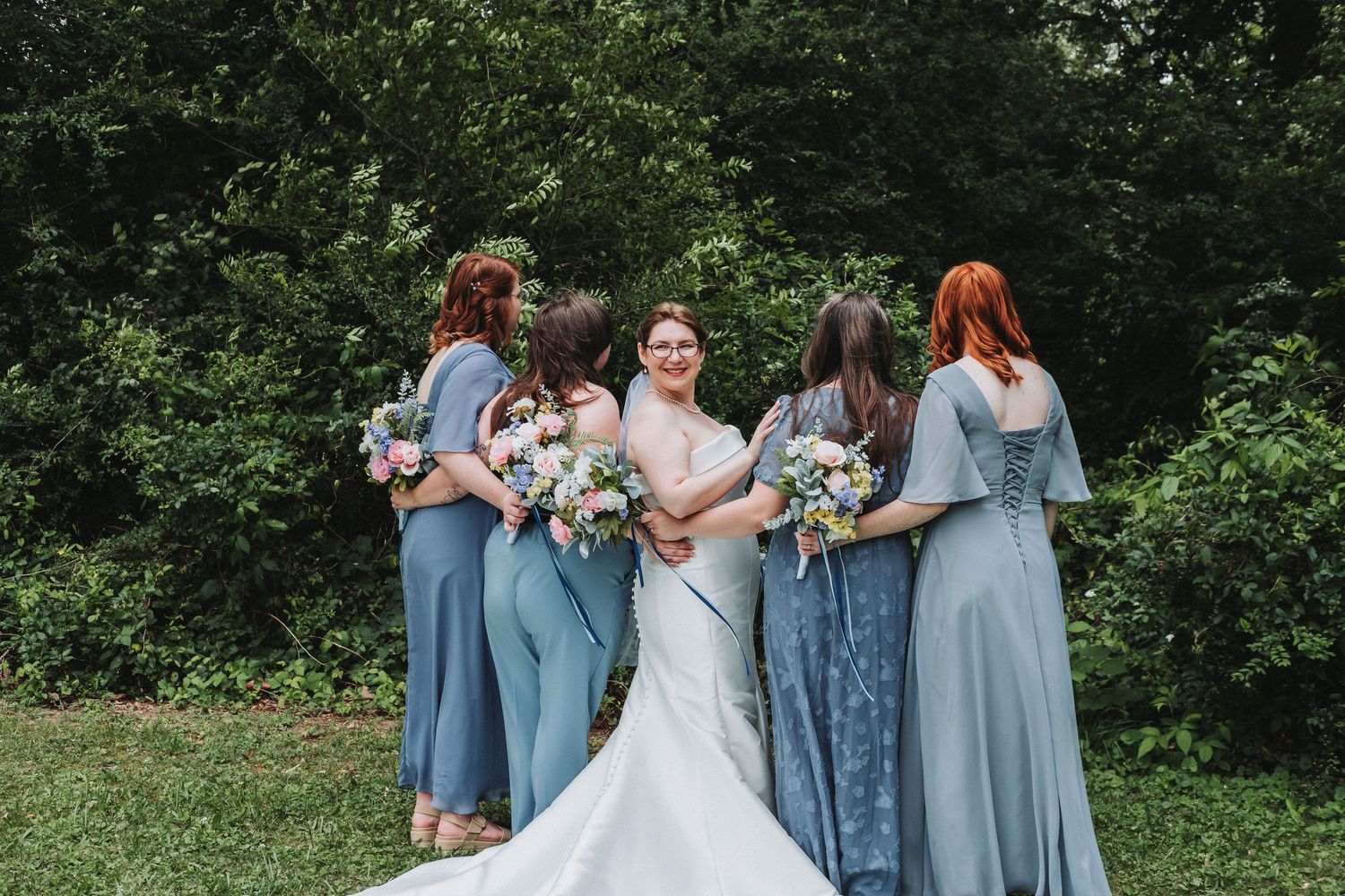 A line of bridesmaids in light blue dresses stand in a row with their backs to camera in an outdoor garden setting.