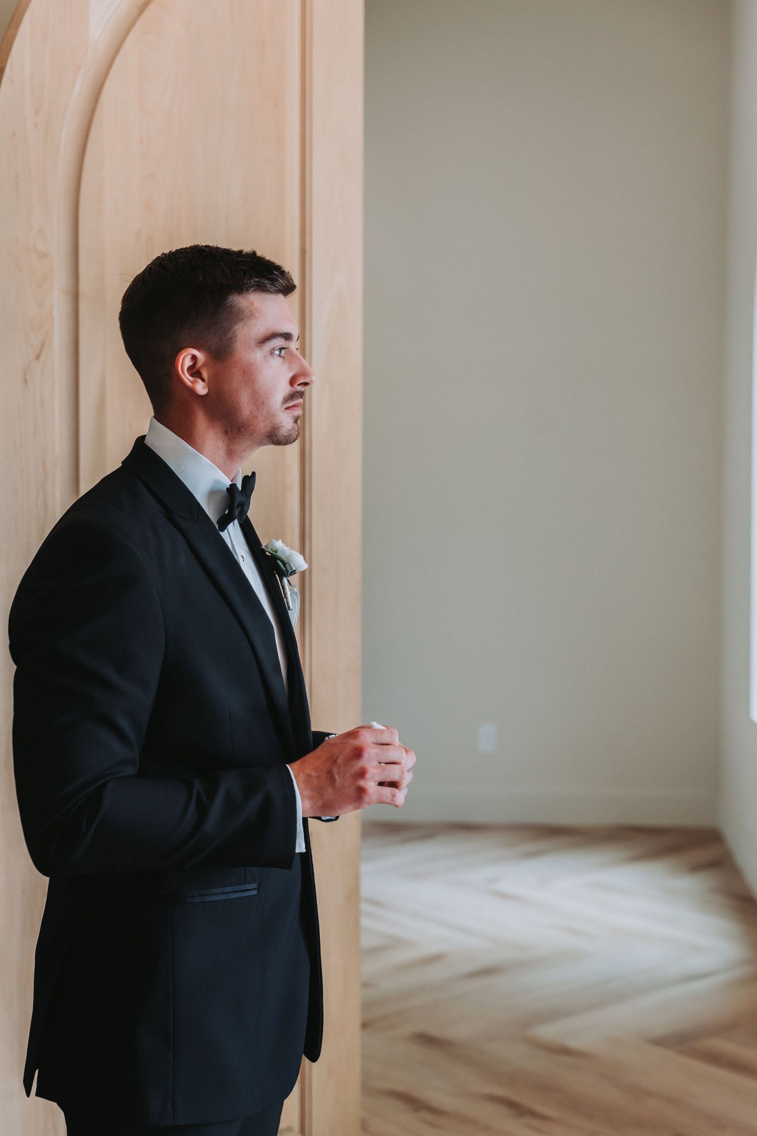 A groom in a classic black tuxedo and bowtie stands pensively by an archway before his wedding ceremony.