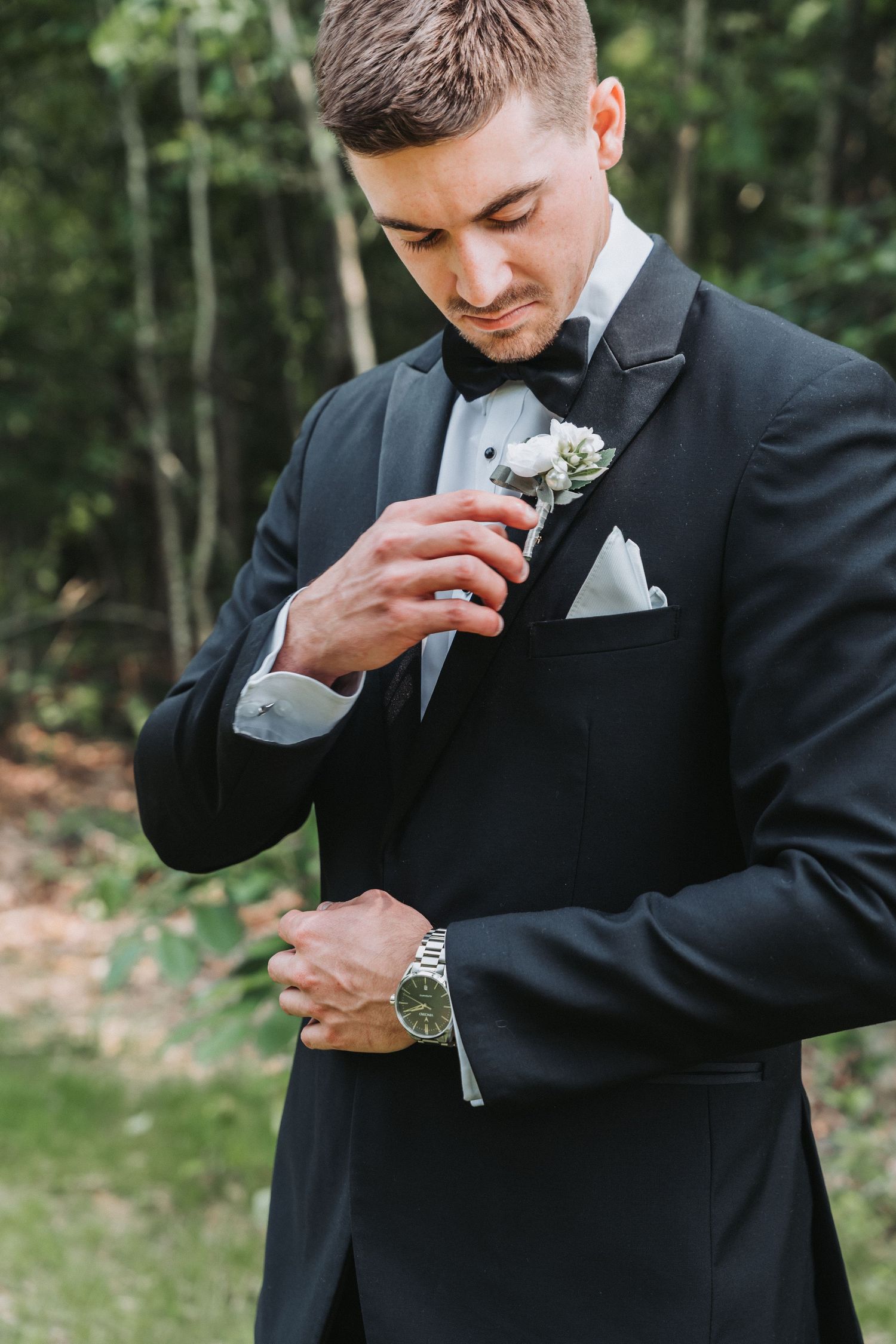 Groom in black tuxedo smells boutonniere flower pinned to his lapel in an outdoor garden setting.