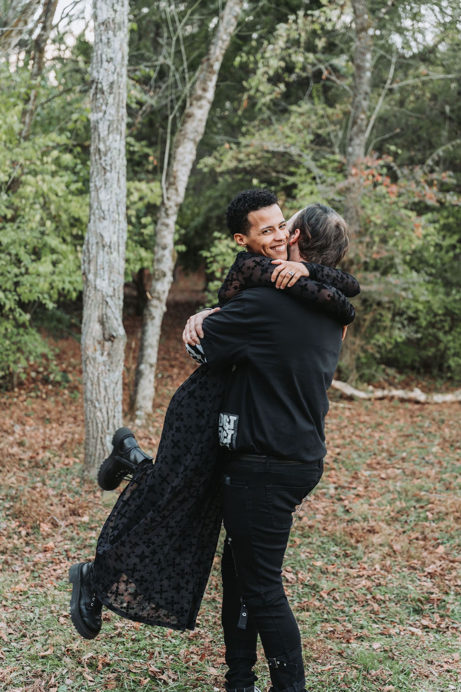 Couple in black outfits embracing in a forest, man lifting woman as they share a romantic moment outdoors.