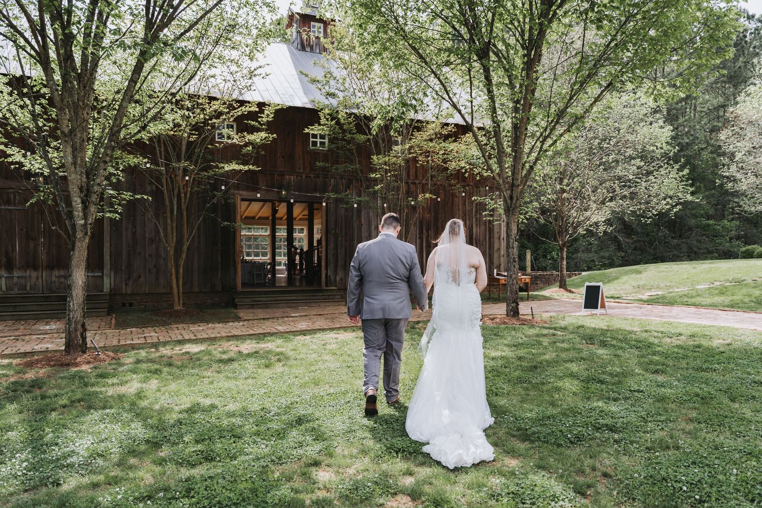 A couple in wedding attire walking together on a green lawn toward a rustic wooden pavilion on a spring day.