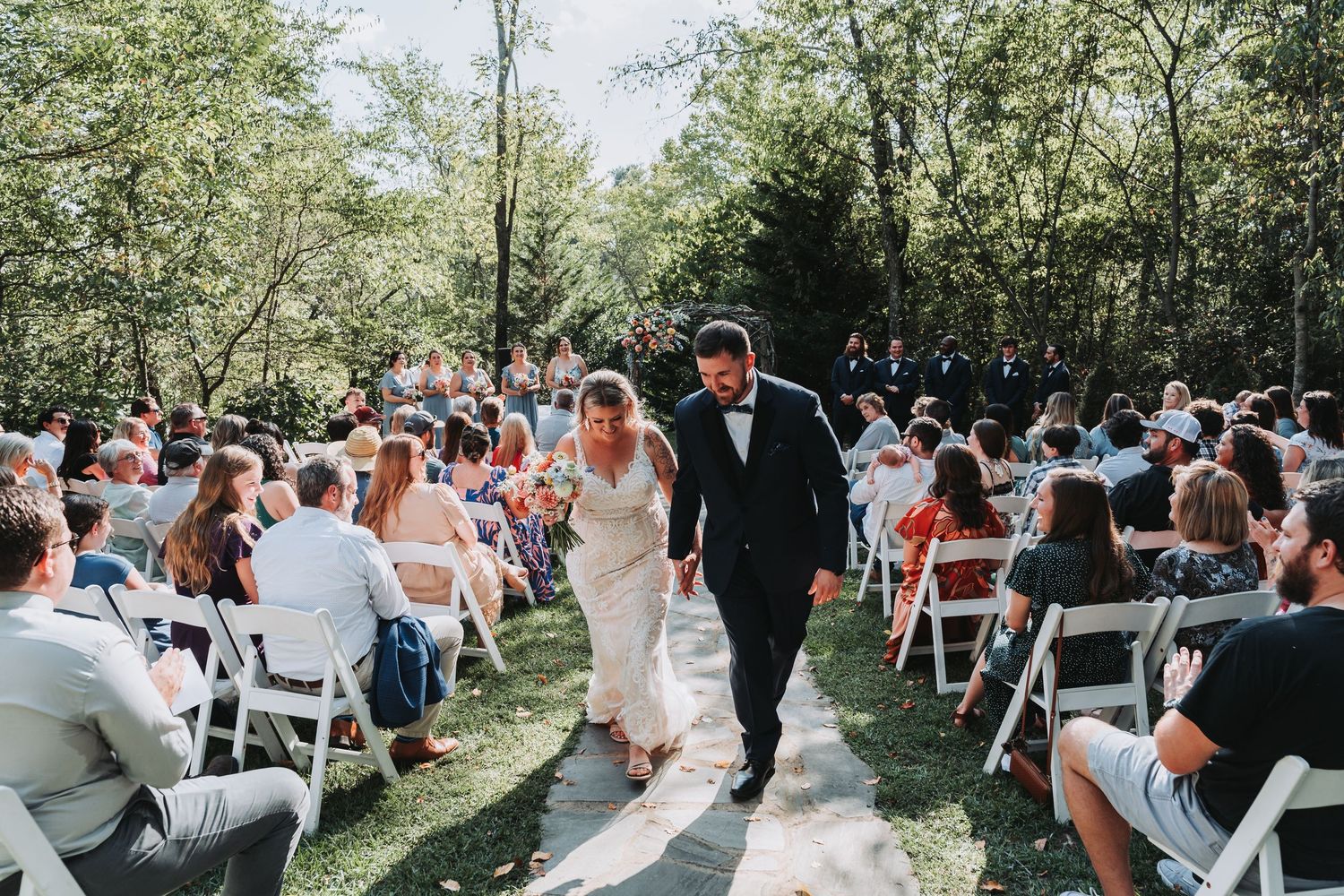 Outdoor wedding ceremony in a wooded area with guests seated on white chairs watching the couple exchange vows.