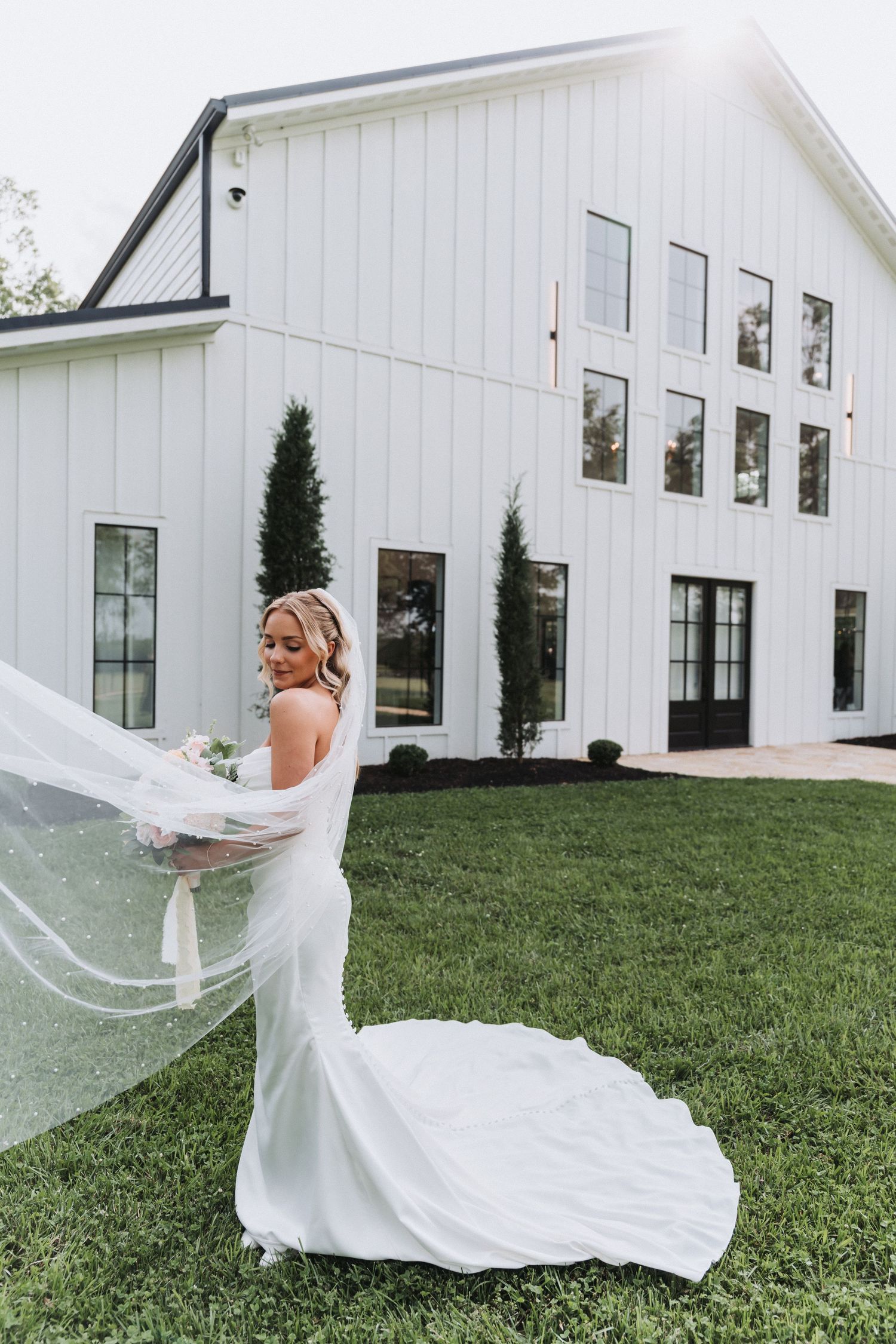 A modern white farmhouse wedding venue with flowing veil and elegant bridal gown on the lawn.