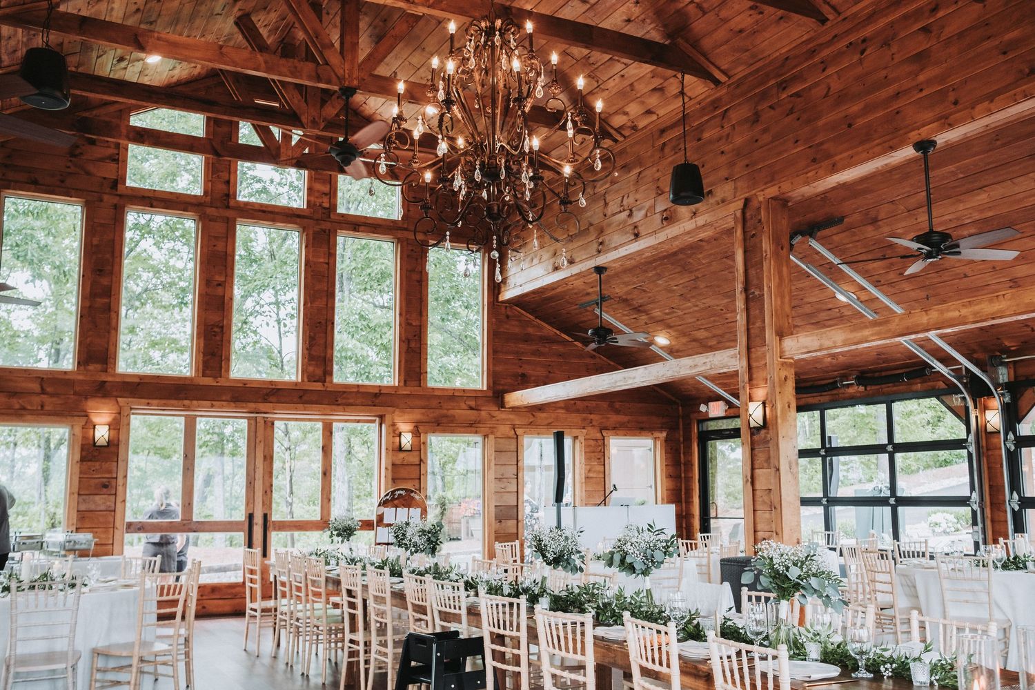 Rustic wooden event venue with exposed beams, chandeliers, and white chairs set up for an elegant wedding ceremony.