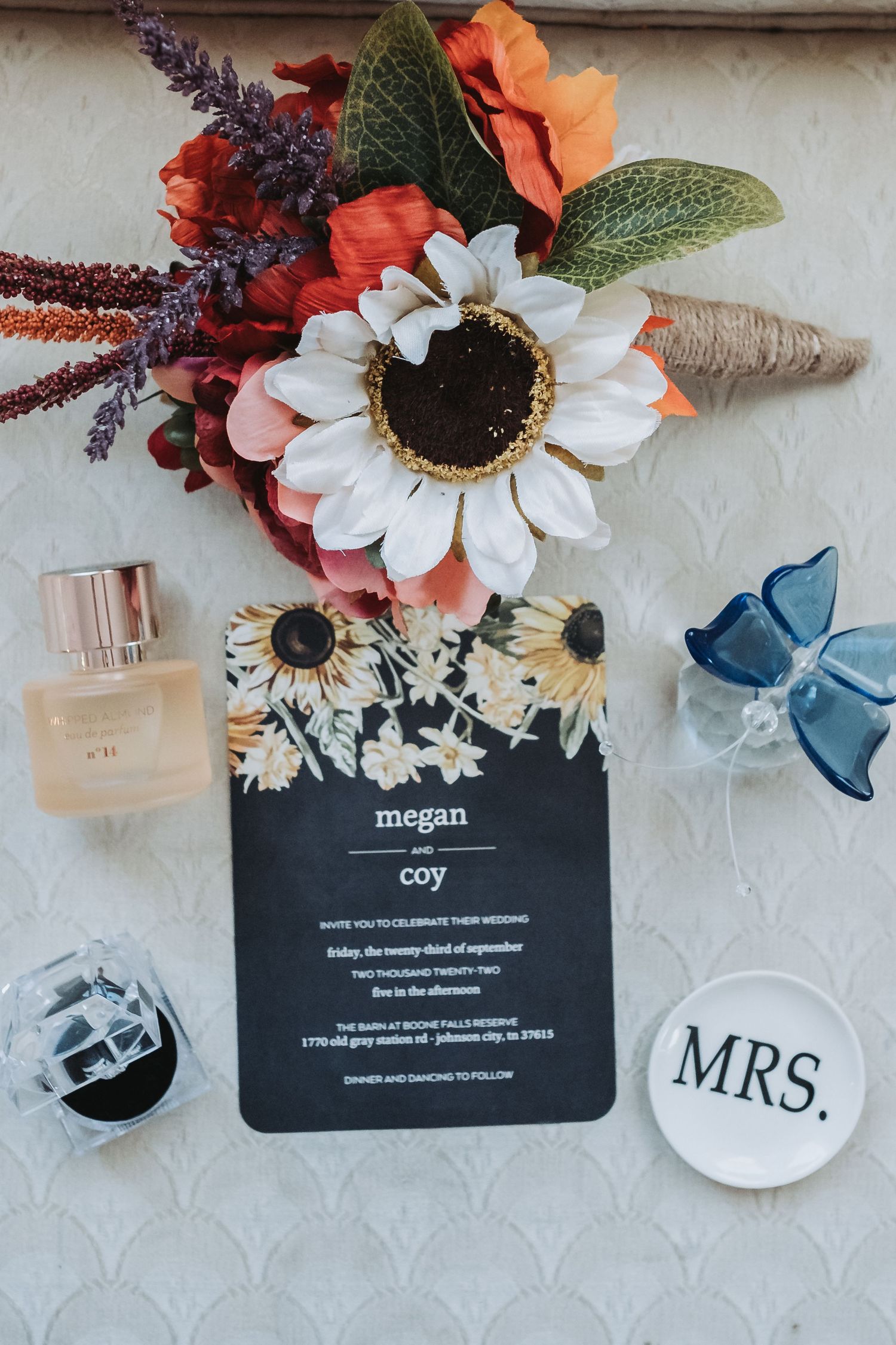 Wedding details flatlay with orange flowers, black invitation card, perfume bottle, MRS sign and blue bow against white background.