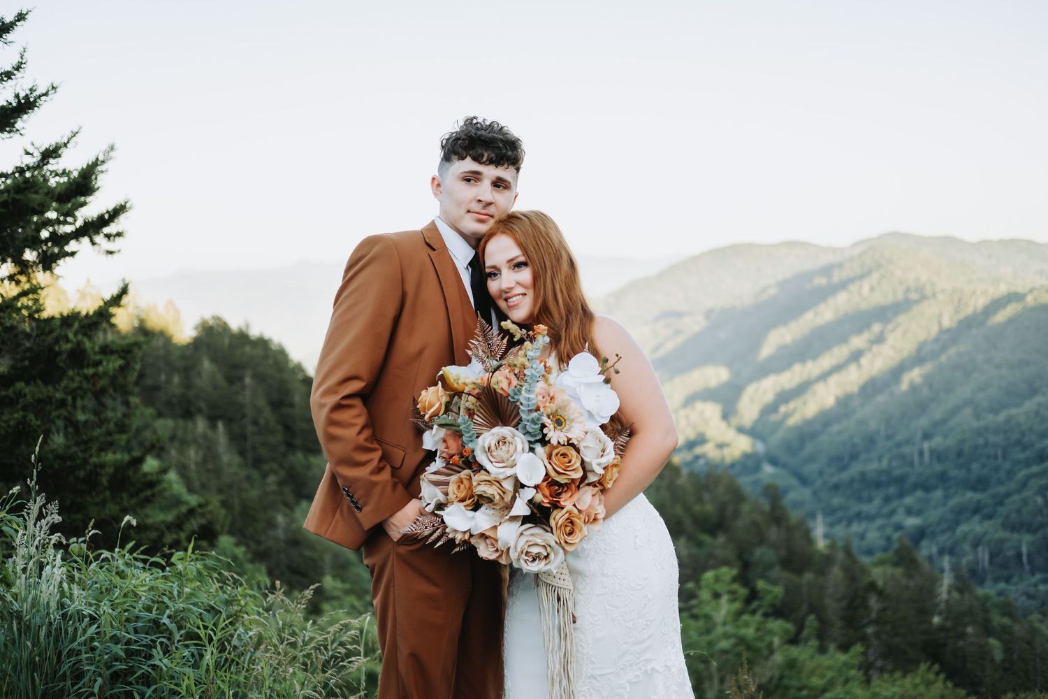 A romantic mountain wedding photo with a brown suit and white dress against a scenic forest backdrop.