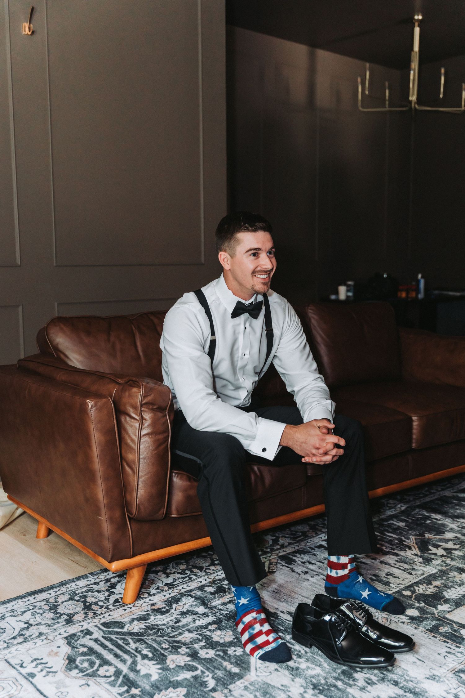 A pair of black dress shoes with colorful patterned socks visible as someone sits on a brown leather couch.